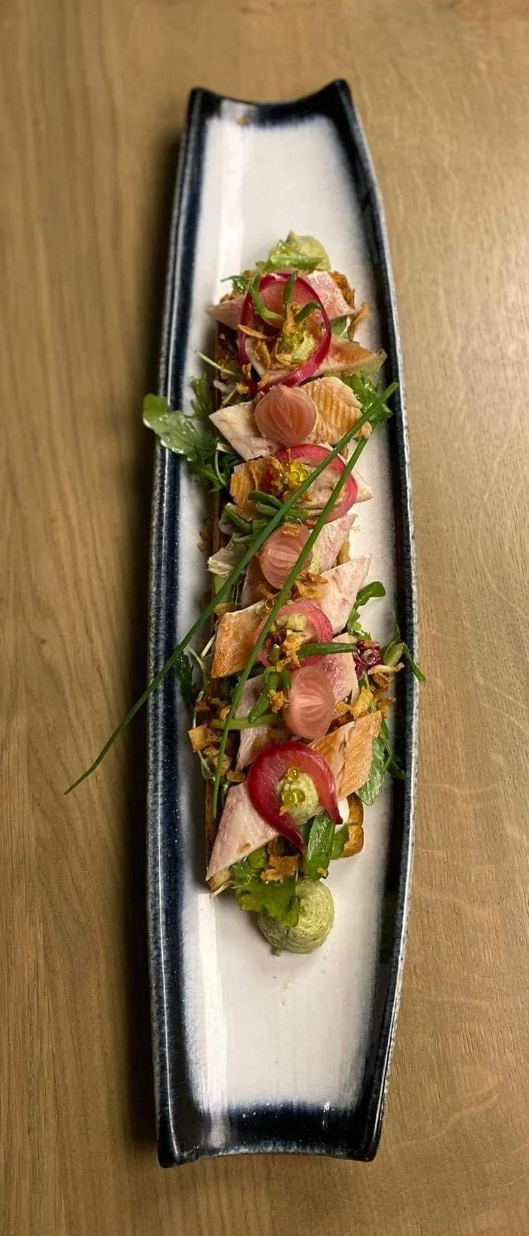Salade artistique avec du poisson, des radis, des herbes et une sauce sur une longue assiette en céramique noire et blanche