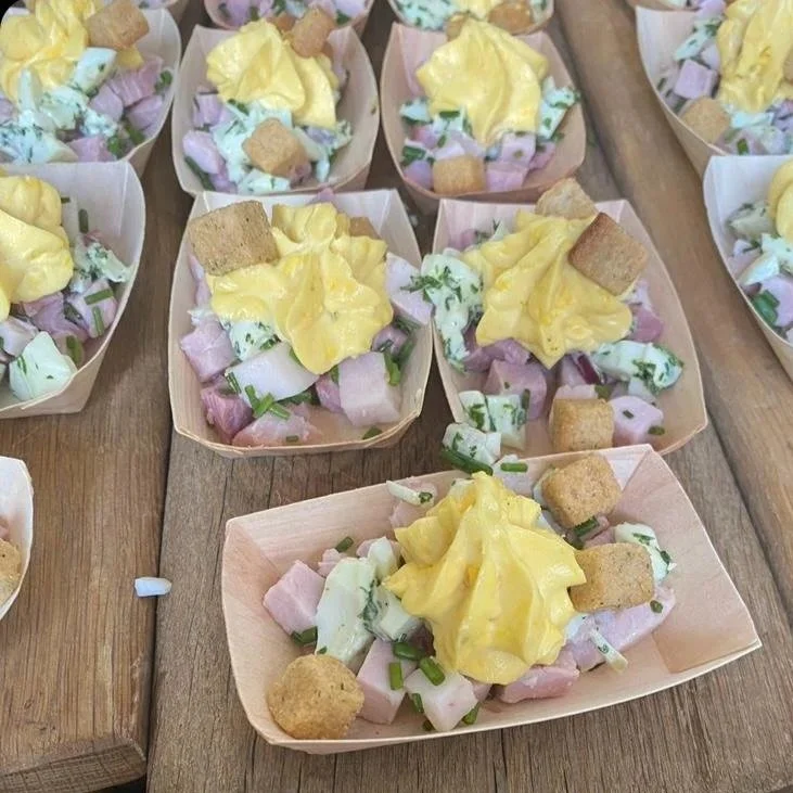 Petites portions de salade servies dans des barquettes en papier, garnies de mayonnaise, cubes de jambon, de concombre, de chaises croutons, sur une surface en bois.