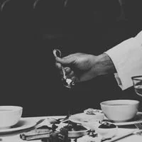 Main d'une personne en costume tenant un verre, avec des assiettes et des tasses sur une table.