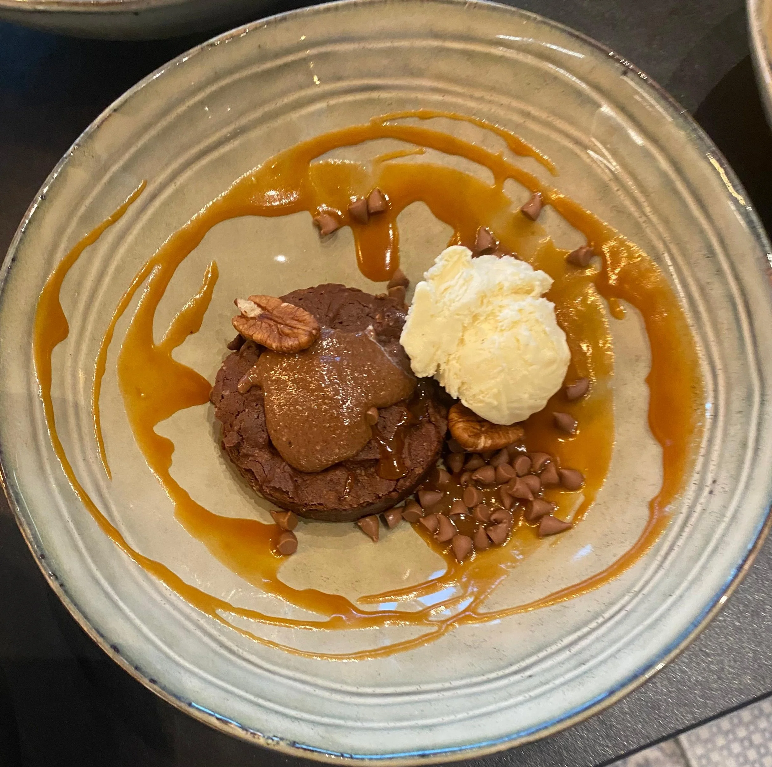 Un plat présenté dans une assiette, comprenant un gâteau au chocolat, une boule de glace à la vanille, des morceaux de noix et des pépites de chocolat, avec un caramel décoratif autour.