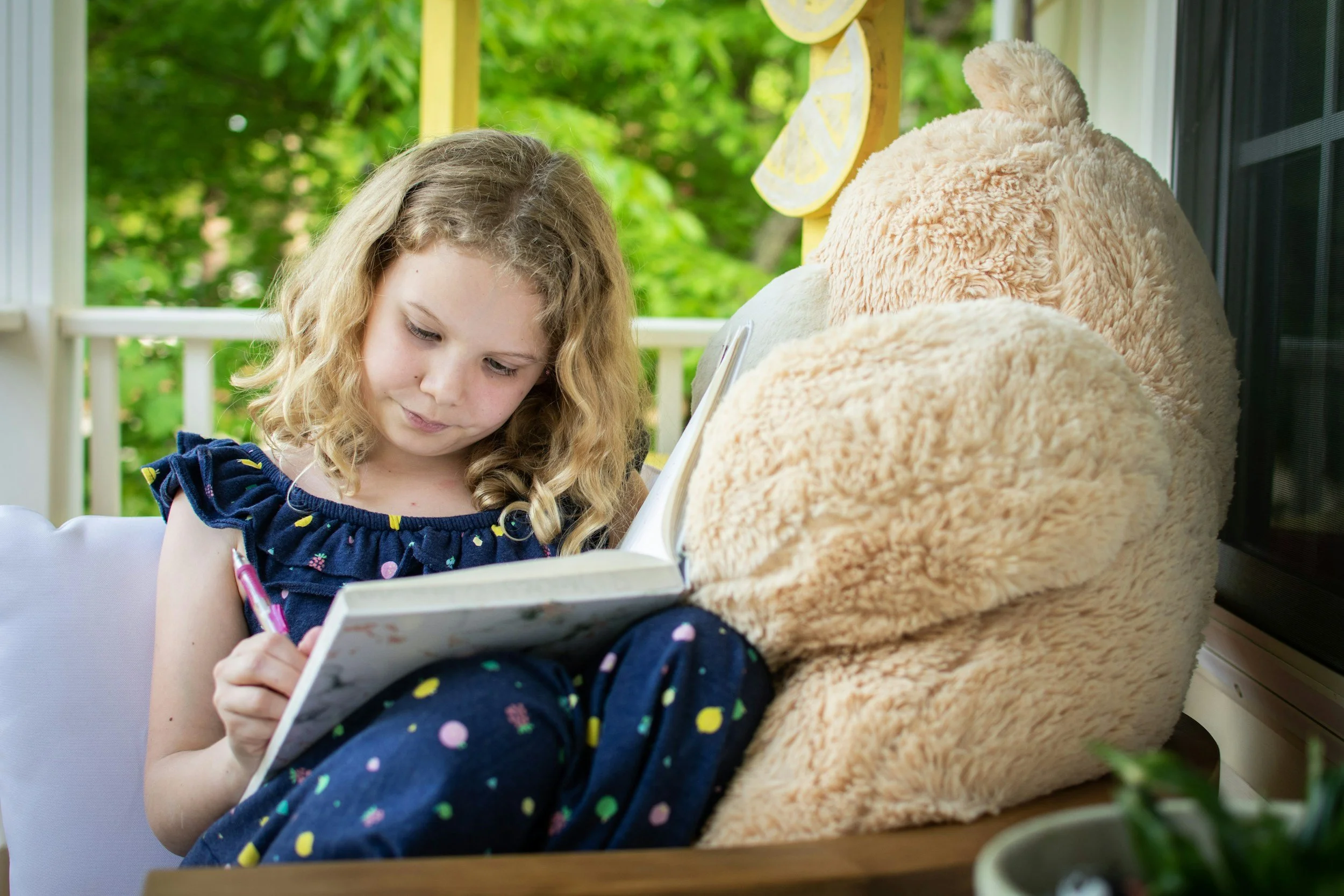 A young girl with curly blonde hair reading a book on a porch, sitting next to a large, plush teddy bear. The porch has a yellow decorative element with lemon slices and greenery in the background.