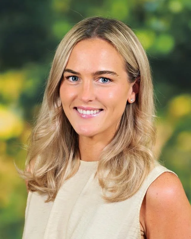 A young woman with blonde hair, blue eyes, and a bright smile, wearing a sleeveless beige top, standing outdoors with a blurred green and yellow natural background.