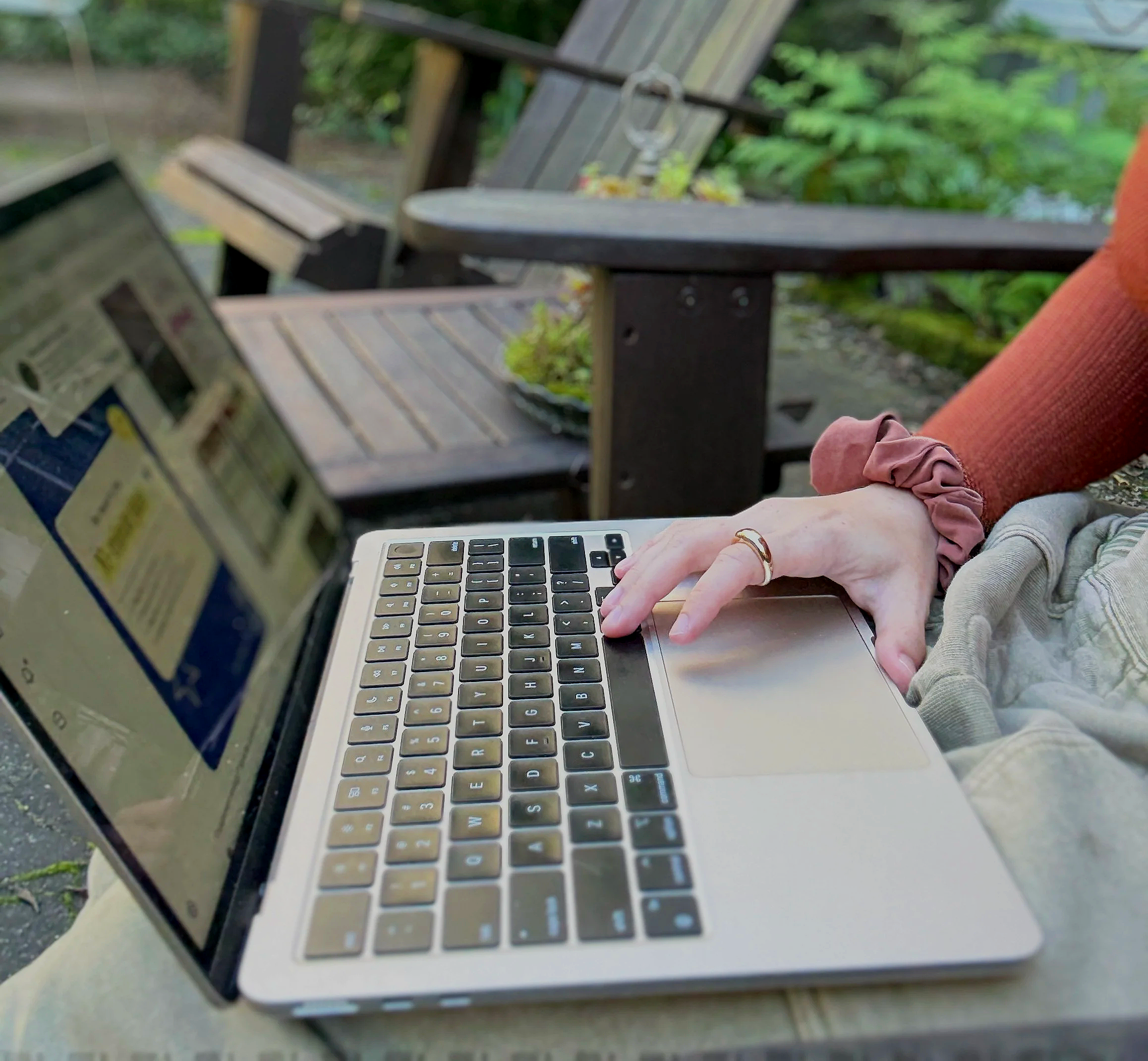 A person sitting outdoors at a wooden table using a silver laptop with a black keyboard.