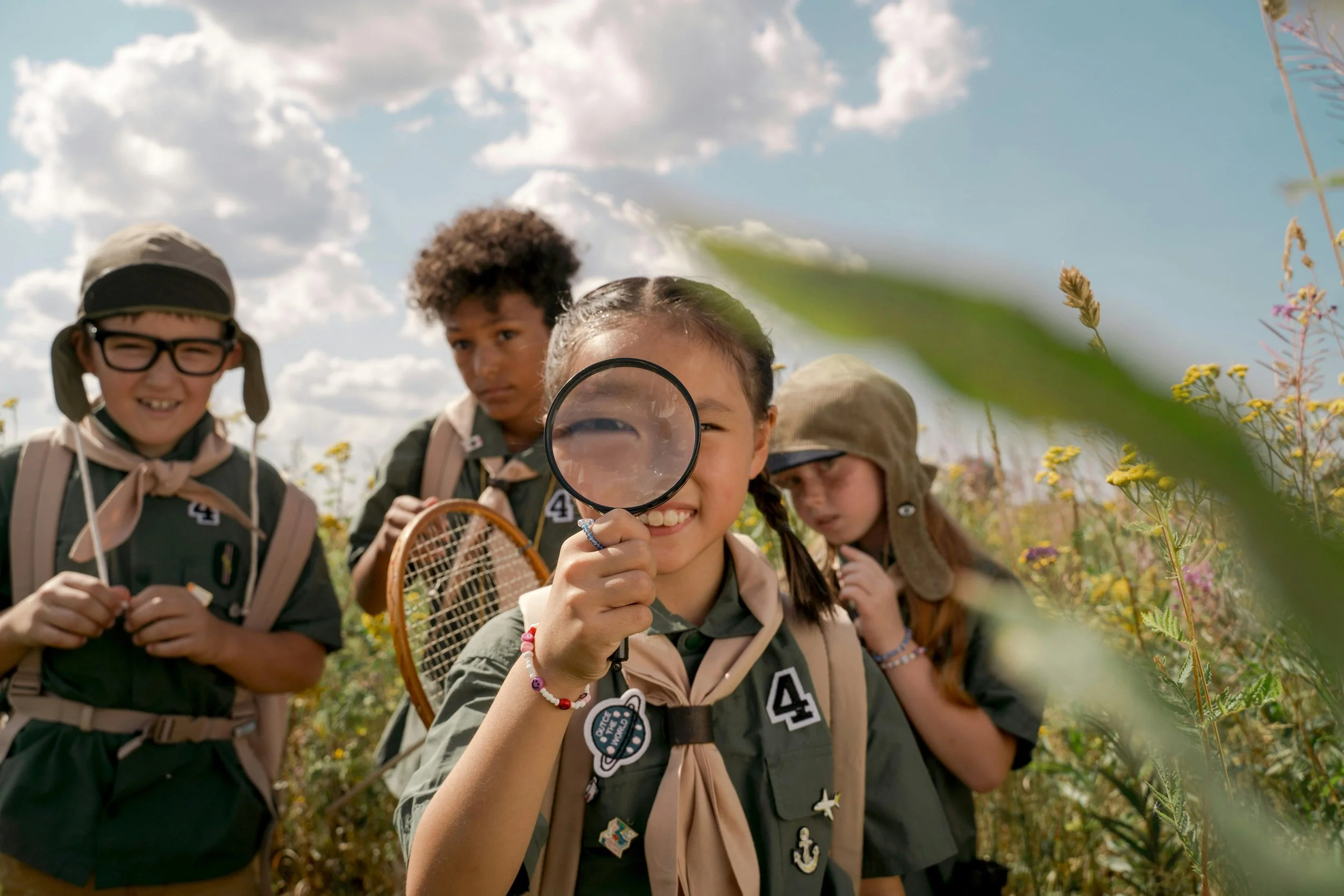 Group of children outdoors, with one girl holding a magnifying glass and inspecting plants, surrounded by other children with backpacks, under a partly cloudy sky.