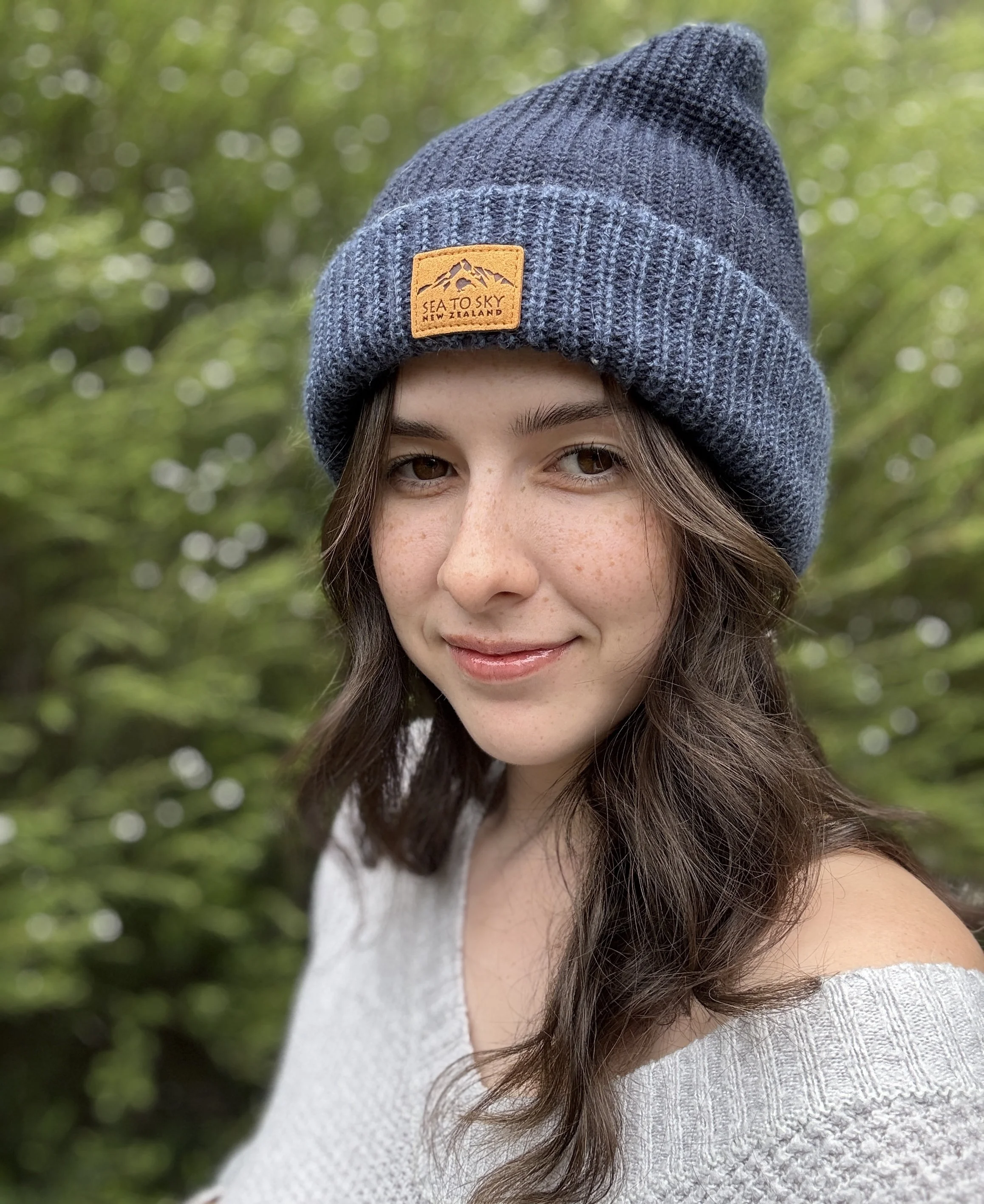 A young woman with long brown hair wearing a gray knit beanie and a white off-shoulder sweater outdoors with a blurred green foliage background.