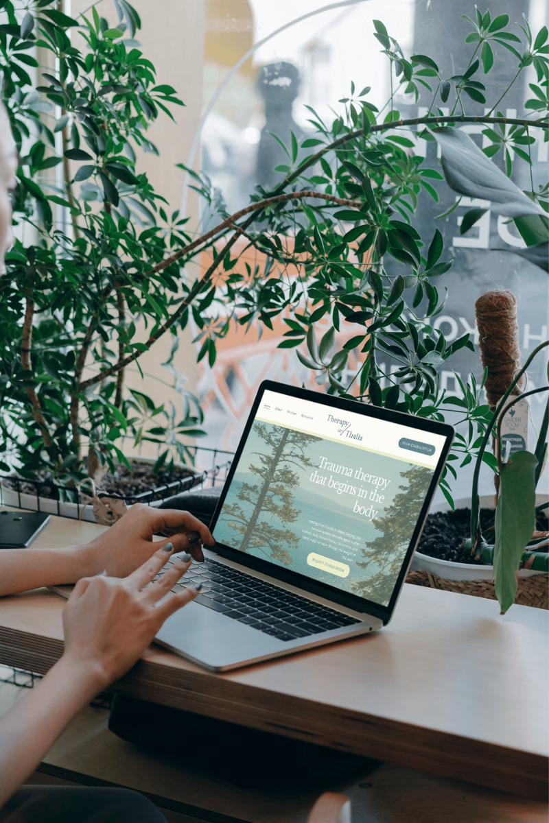 A person is sitting at a wooden table, using a laptop that displays a website about trauma therapy with trees in the background. There are large green plants surrounding the workspace near a window.