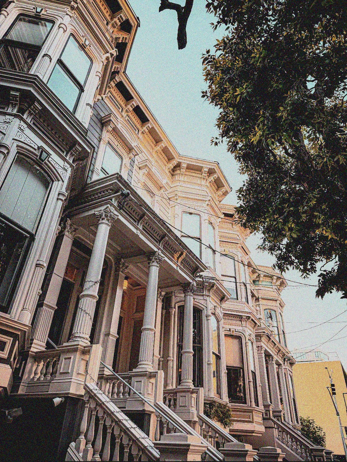 A row of Victorian-style houses with intricate architectural details, large windows, and staircases leading up to front doors, with green trees and power lines nearby.