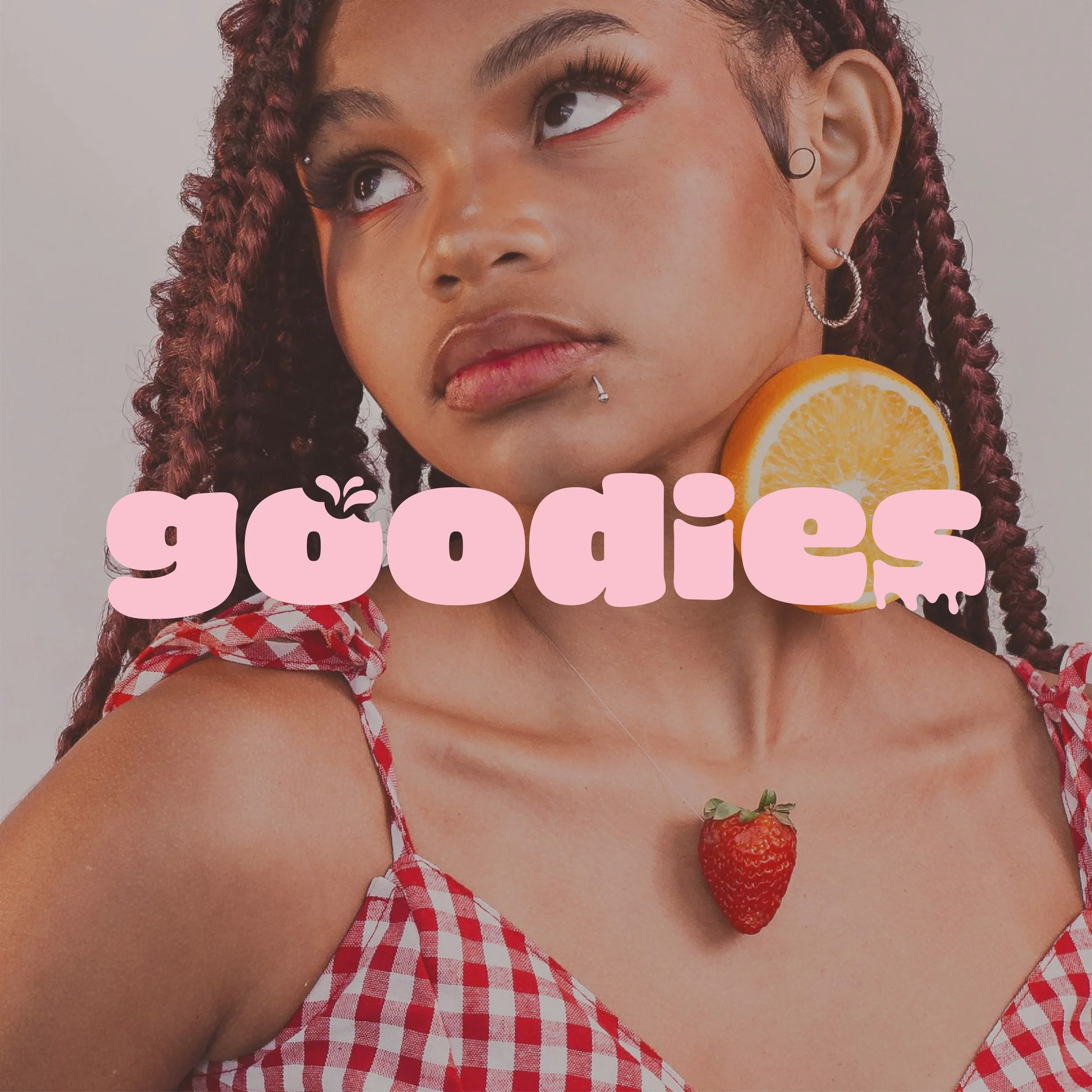 Close-up of a woman with braided hair, wearing earrings, a red checkered top, with strawberries and an orange slice as accessories, and pink text overlay.