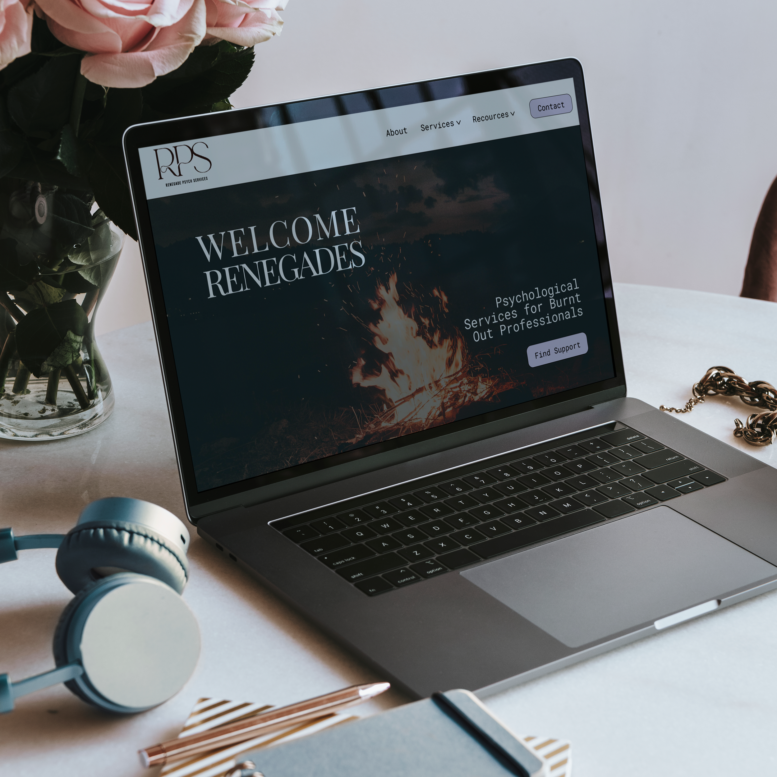 Laptop displaying a website titled "WELCOME RENEGADES" with a dark background and a light menu bar at the top, surrounded by pink roses in a vase on the left, and gold jewelry, a pair of over-ear headphones, striped straws, and a roll of tape on a white surface.