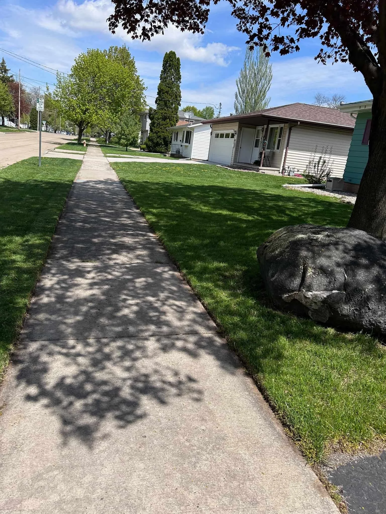 A sidewalk runs through a residential neighborhood with green lawns, trees, and houses on a sunny day.