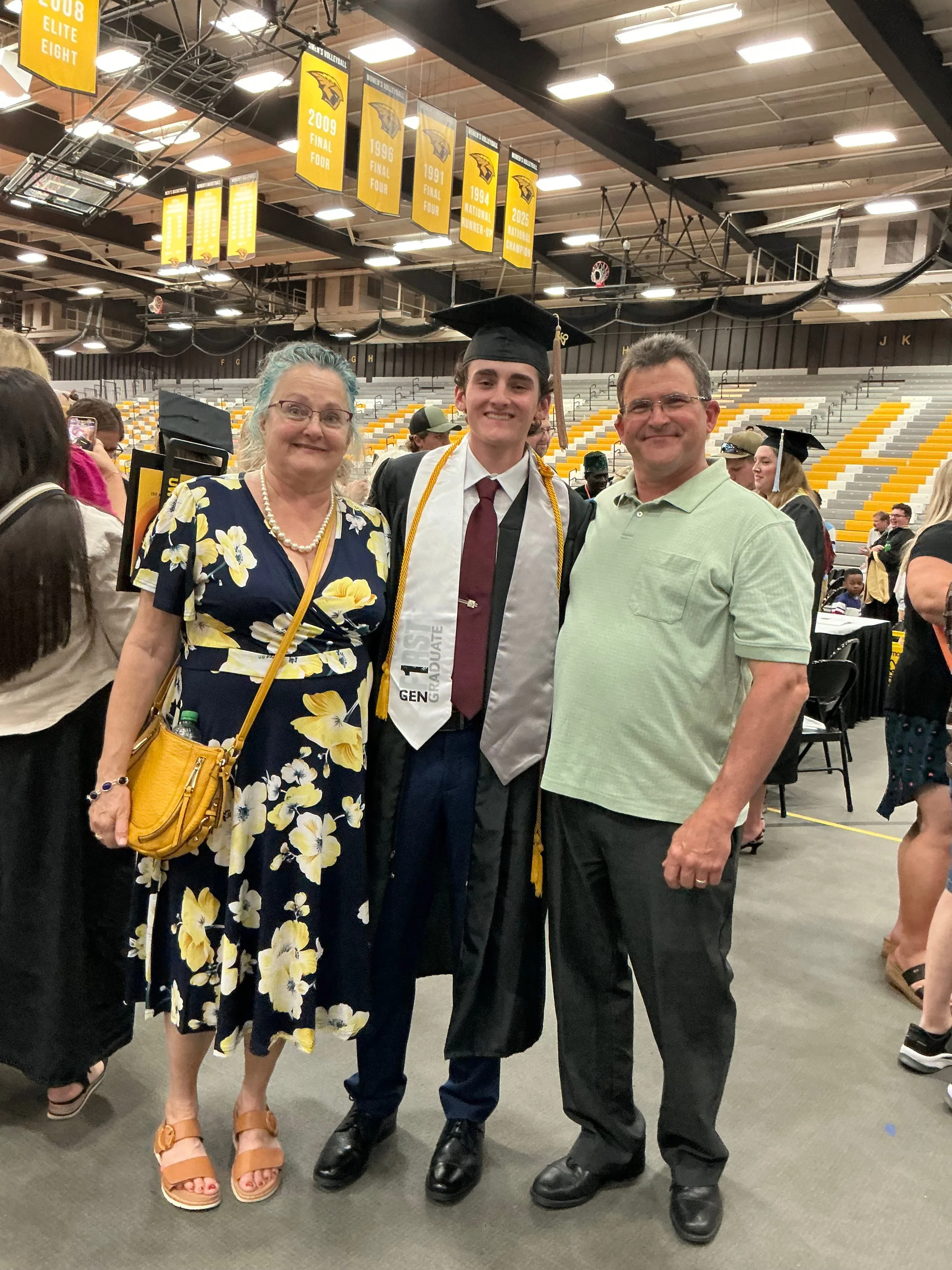 A group of three people standing together at a graduation ceremony inside a gymnasium. The graduate is in the center wearing a black cap and gown with a white sash that says "1st Generation Graduate." They are all smiling and posing for the photograph.