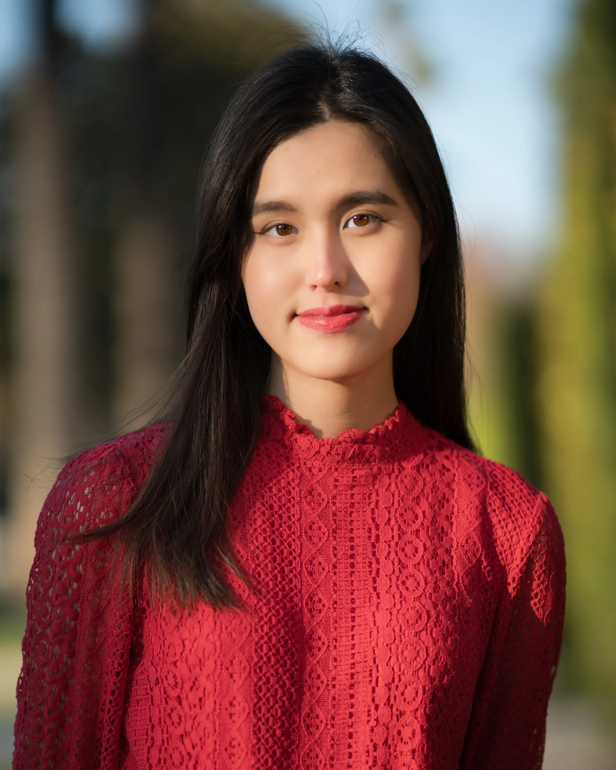 A young woman with long dark hair wearing a red lace dress outdoors, with blurred trees in the background.