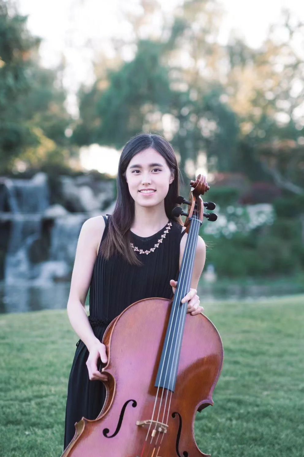Young woman outdoors holding a cello, with trees and water feature in the background.