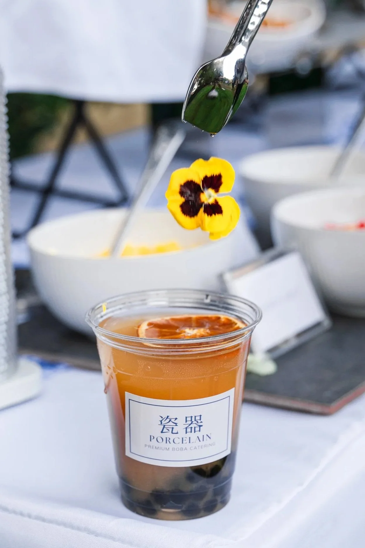 A plastic cup of bubble tea with dark tapioca pearls at the bottom, labeled "Porcelain Premium Boba Catering," with an orange-colored drink and a lemon slice garnish on top. A spoon with a yellow flower is positioned above the cup, with a blurred background of a dining setup.