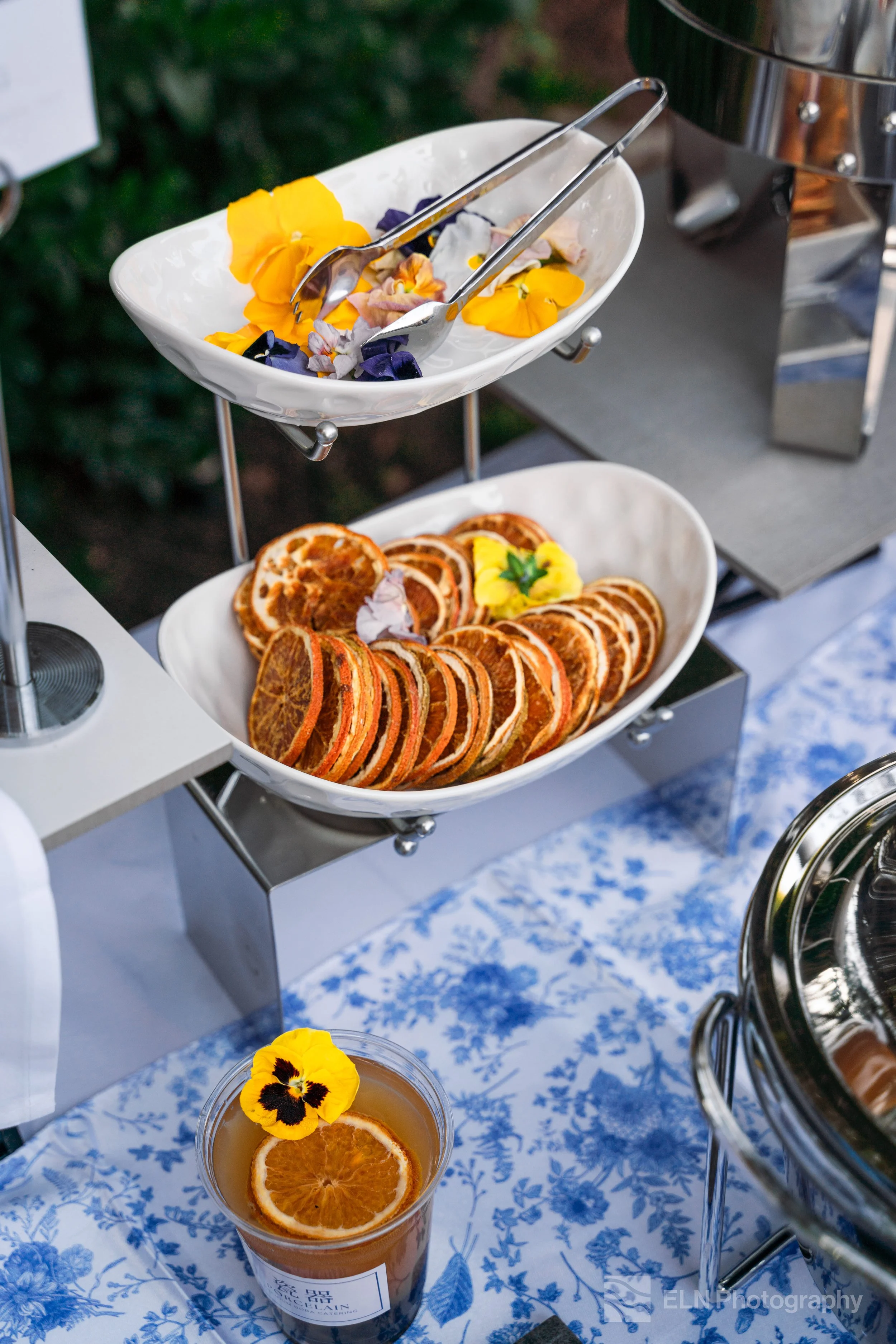 A tiered serving tray with edible flower petals, including yellow and purple flowers, along with sliced oranges and a drink garnished with orange slices and a flower, on a table with a blue and white floral tablecloth.