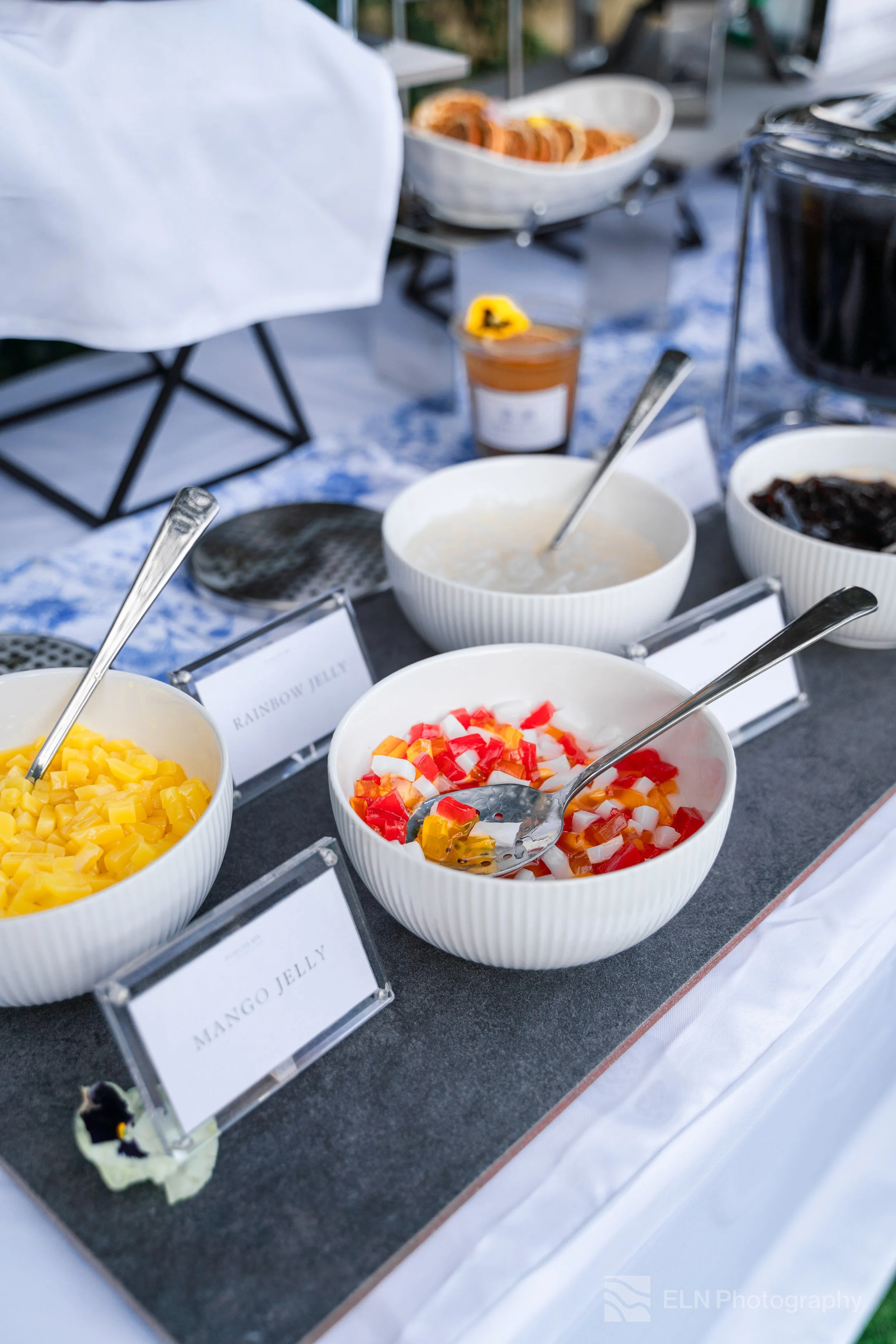 Bowl of diced mango next to a bowl of diced red and white fruit, possibly a fruit salad, with other bowls and food items on a buffet table, with signs indicating mango jello and rainbow jelly.