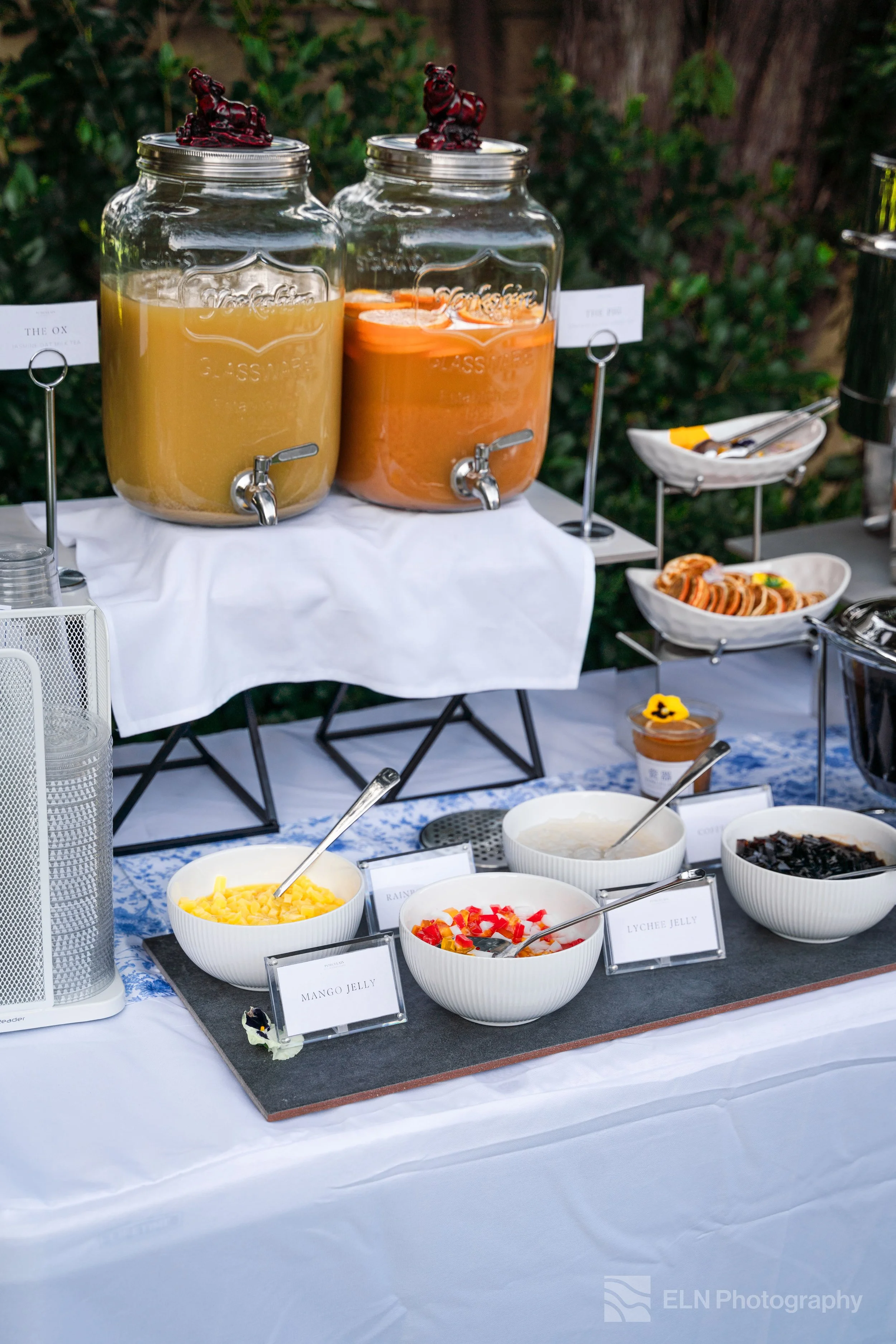 A buffet table with two large glass dispensers filled with orange and yellow drinks on a white tablecloth, with a variety of bowls containing fruit toppings and desserts with labels, set outdoors with greenery in the background.
