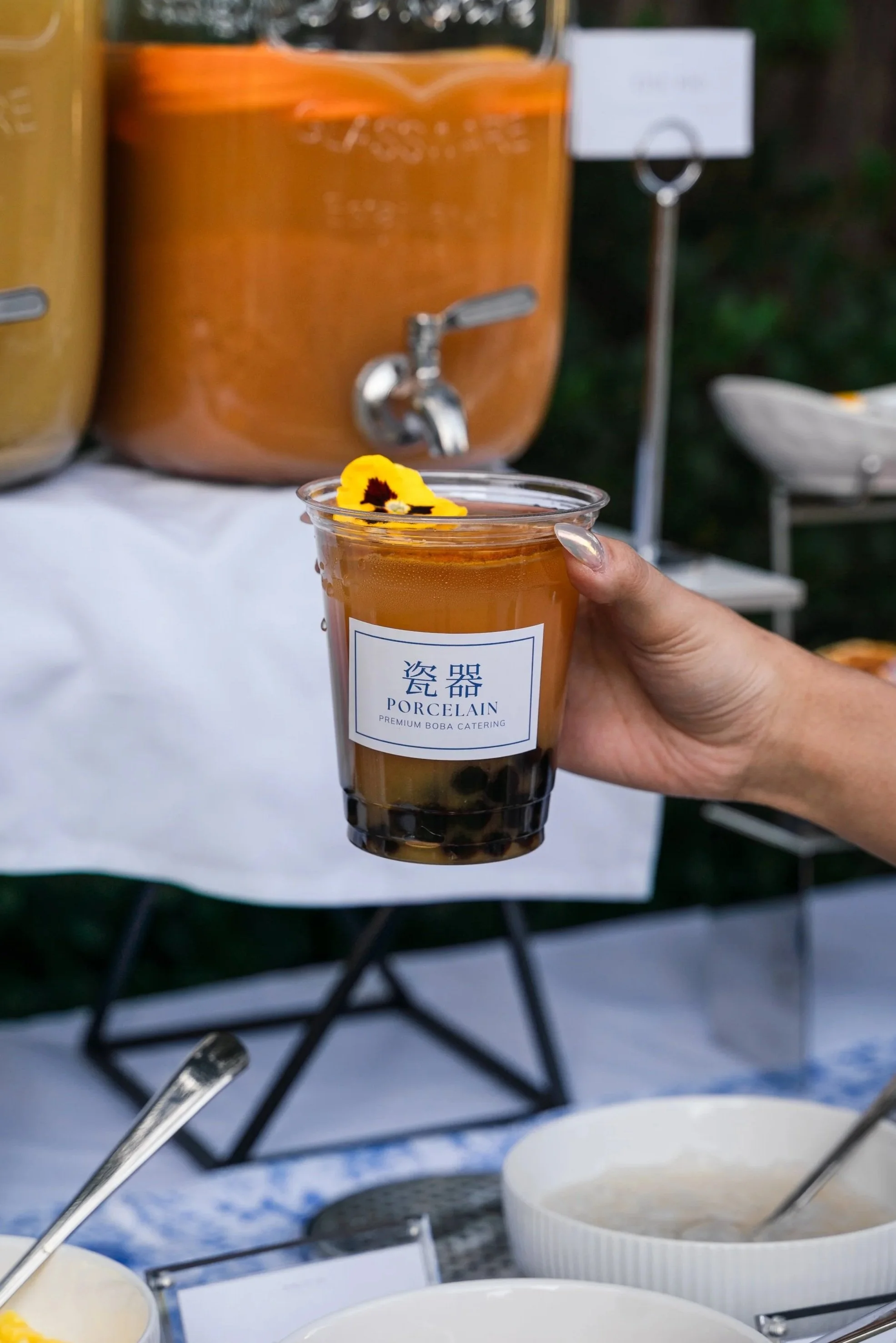 Hand holding Jasmine Oat Milk Tea with edible flower and dried orange slices with branded label.