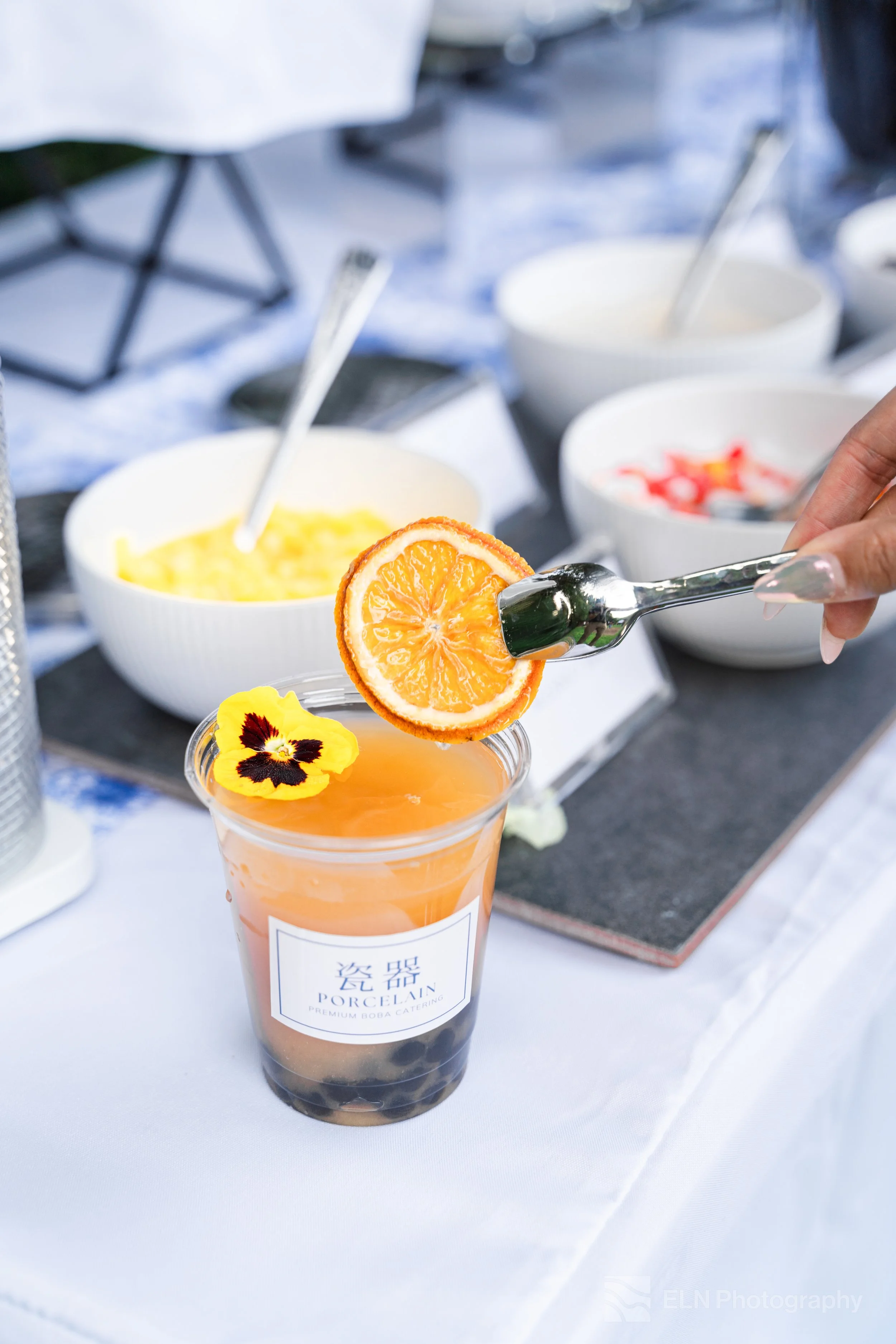 A cup of bubble tea with tapioca pearls and an orange slice garnish, on a table with bowls of toppings in the background.