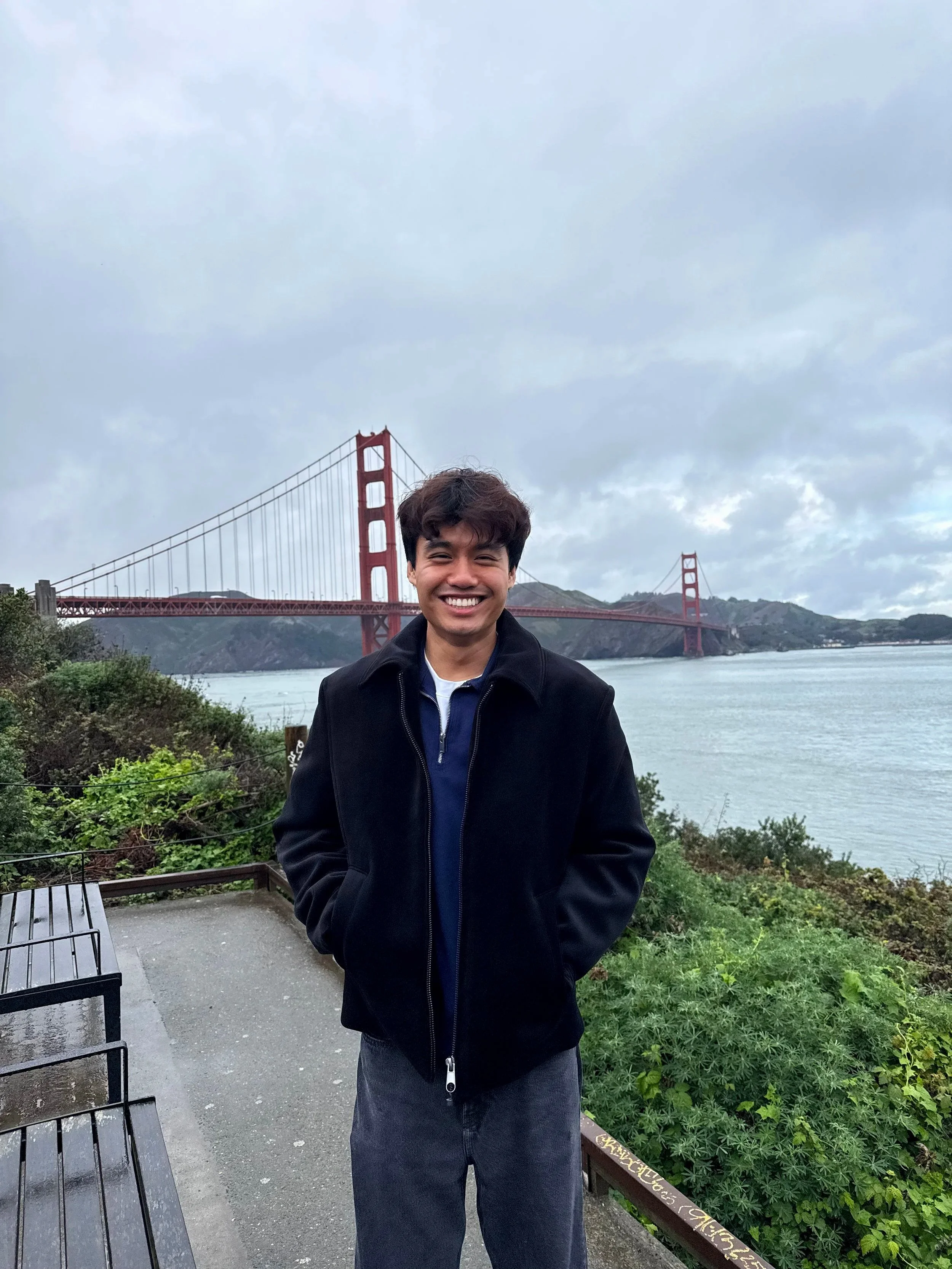 A young man smiling standing outdoors near the Golden Gate Bridge with greenery and a bench nearby, overcast sky in the background.
