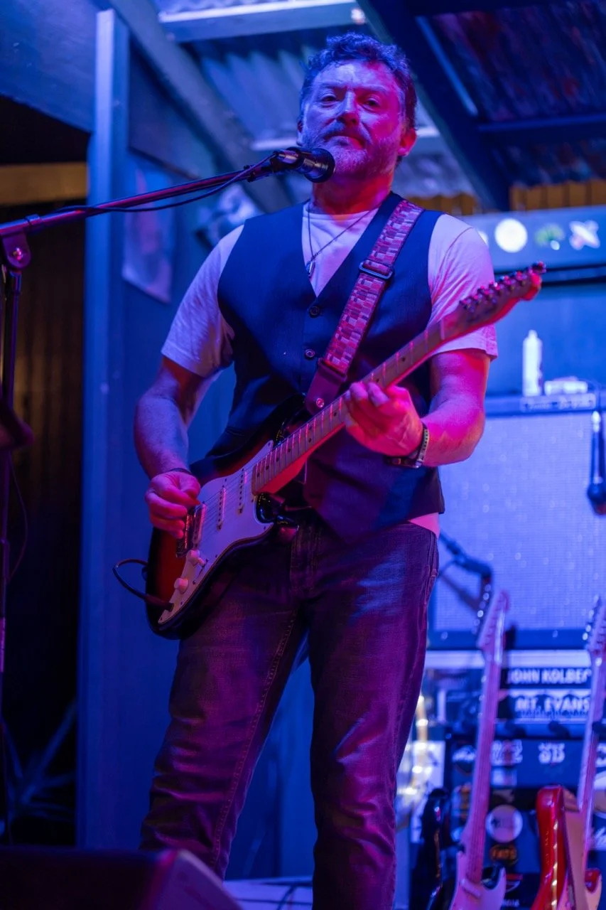 A man playing an electric guitar and singing into a microphone on stage with blue and purple lighting.