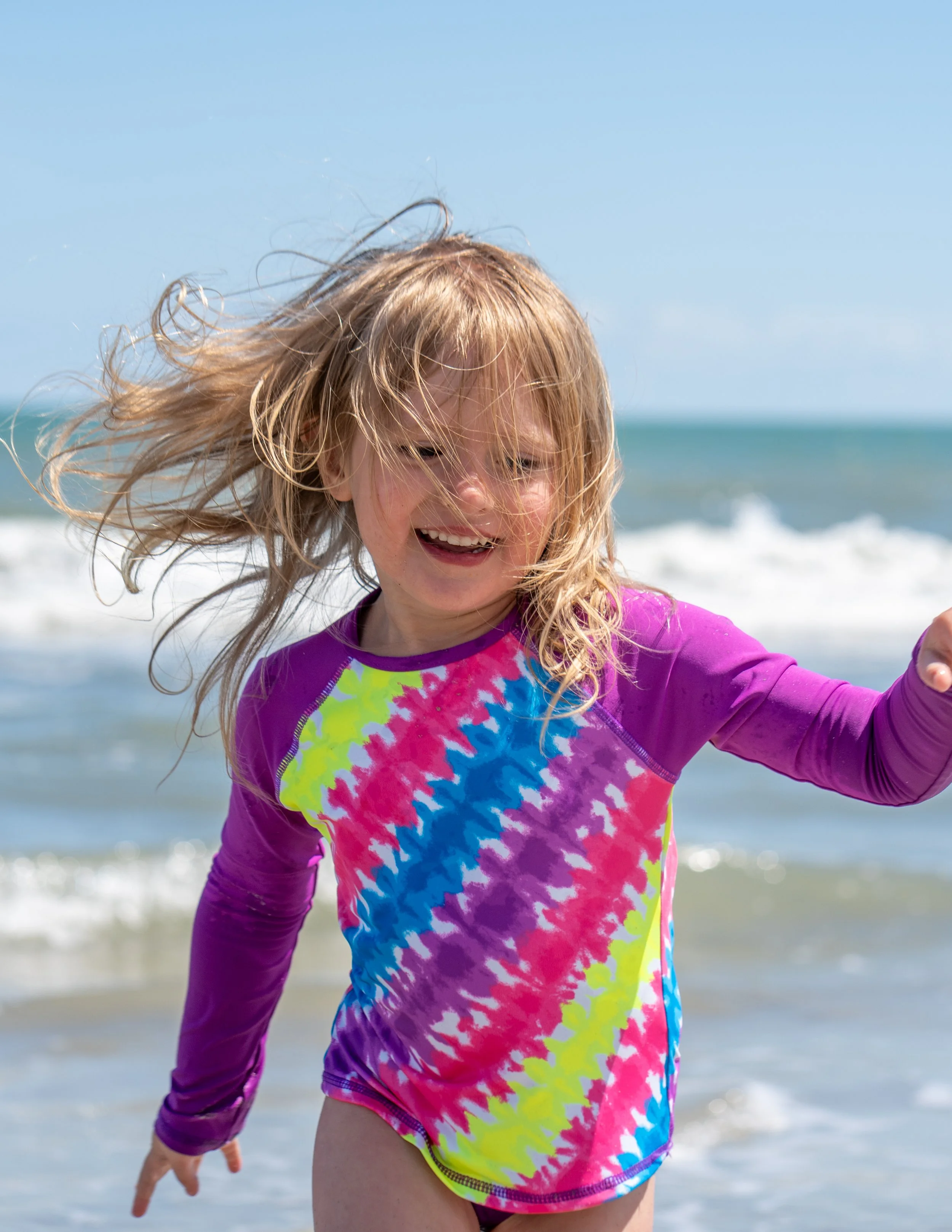 A young girl with long, blonde hair smiling and playing on the beach, wearing a vibrant, tie-dye long-sleeve rash guard with pink, blue, yellow, and green colors. Waves and the ocean are visible in the background.