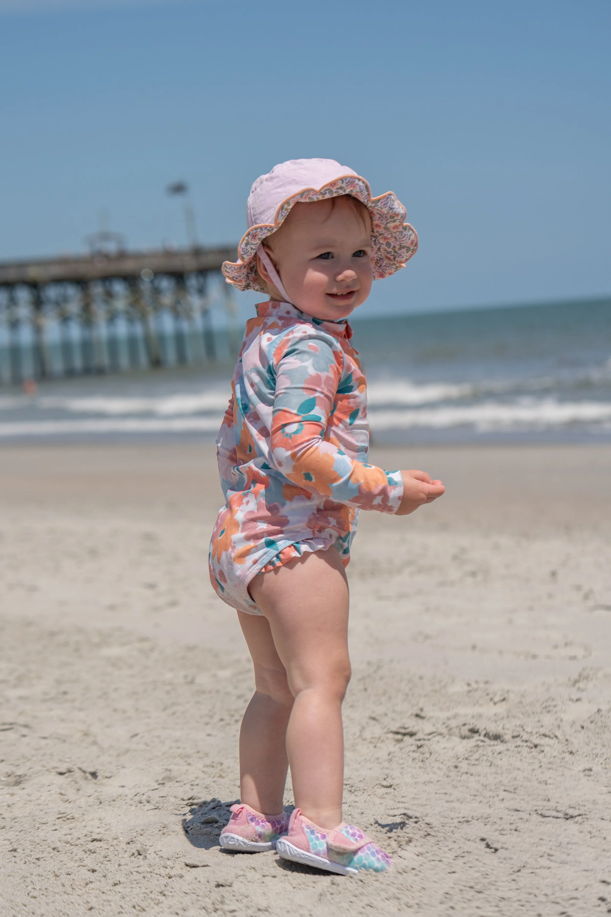 A young girl in a floral rash guard and matching shorts standing on the sandy beach. She is wearing pink sneakers and a pink sun hat. The ocean and a pier with a lighthouse are visible in the background.