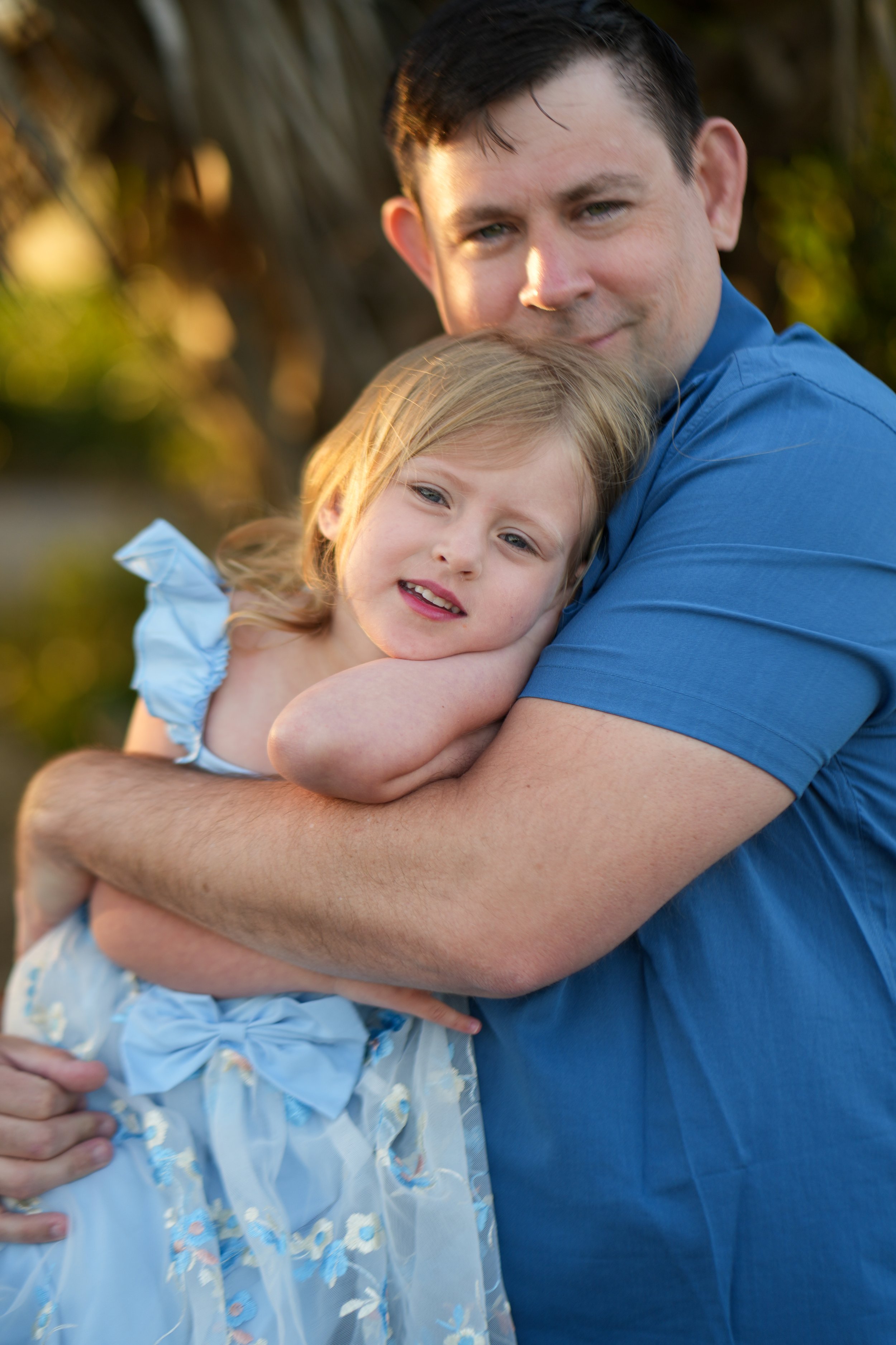 A man with short dark hair and a little girl with blonde hair in a blue dress hug each other outside during the daytime.