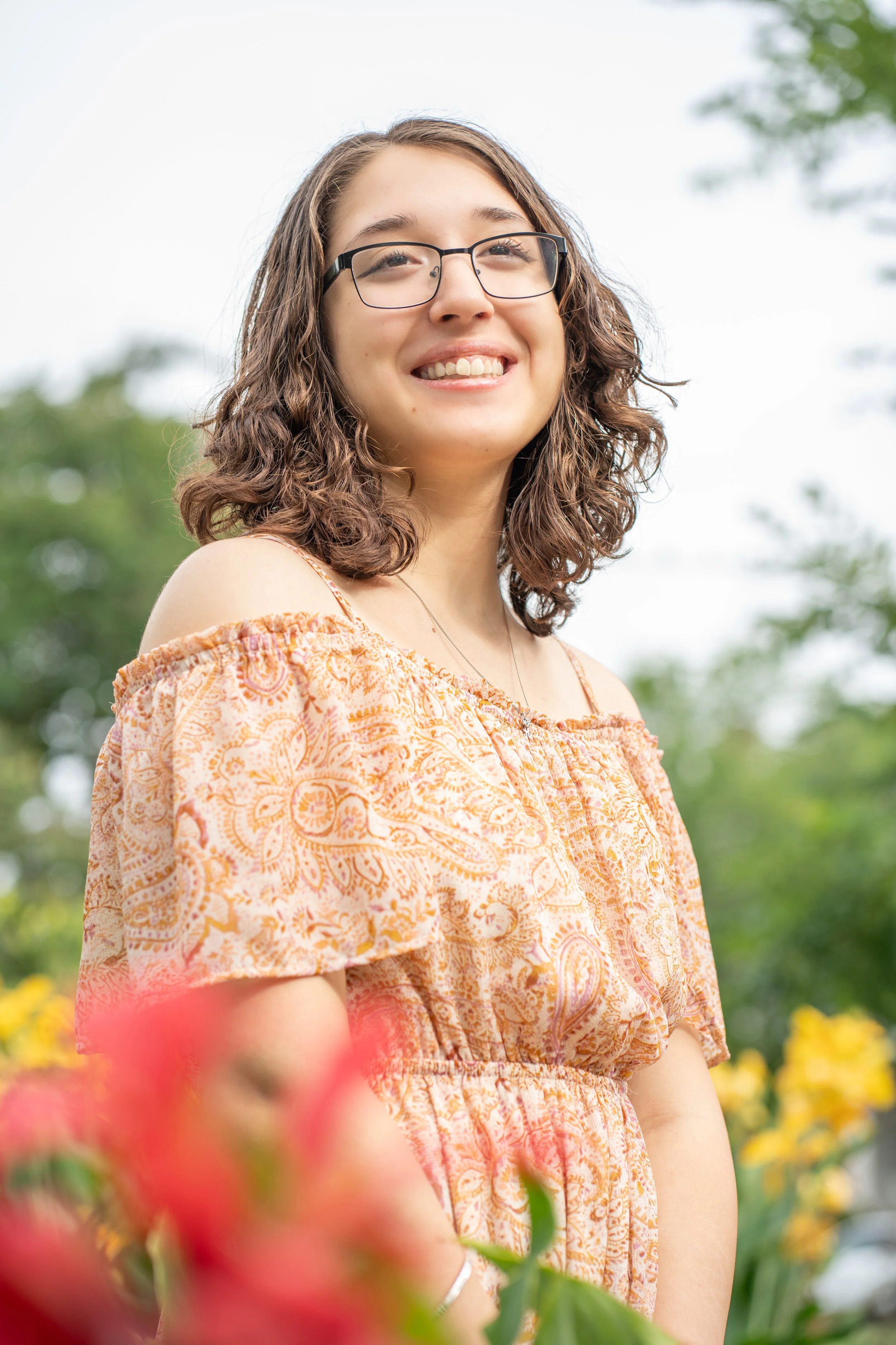 A young woman with curly hair and glasses smiling while standing outdoors in a garden with colorful flowers and trees.