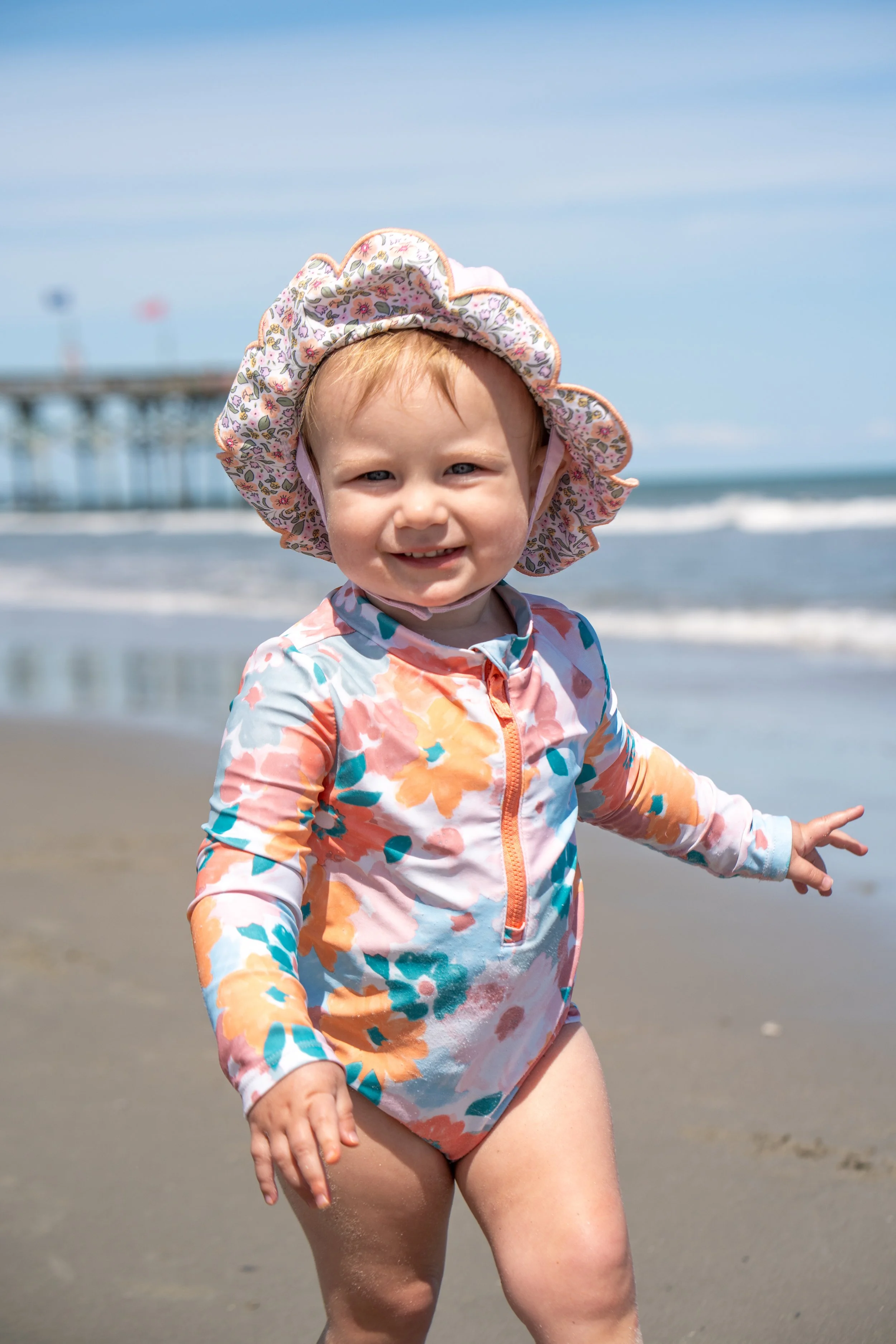 A young girl wearing a floral sunhat and colorful swimsuit is smiling and playing on a beach with waves and a pier in the background.