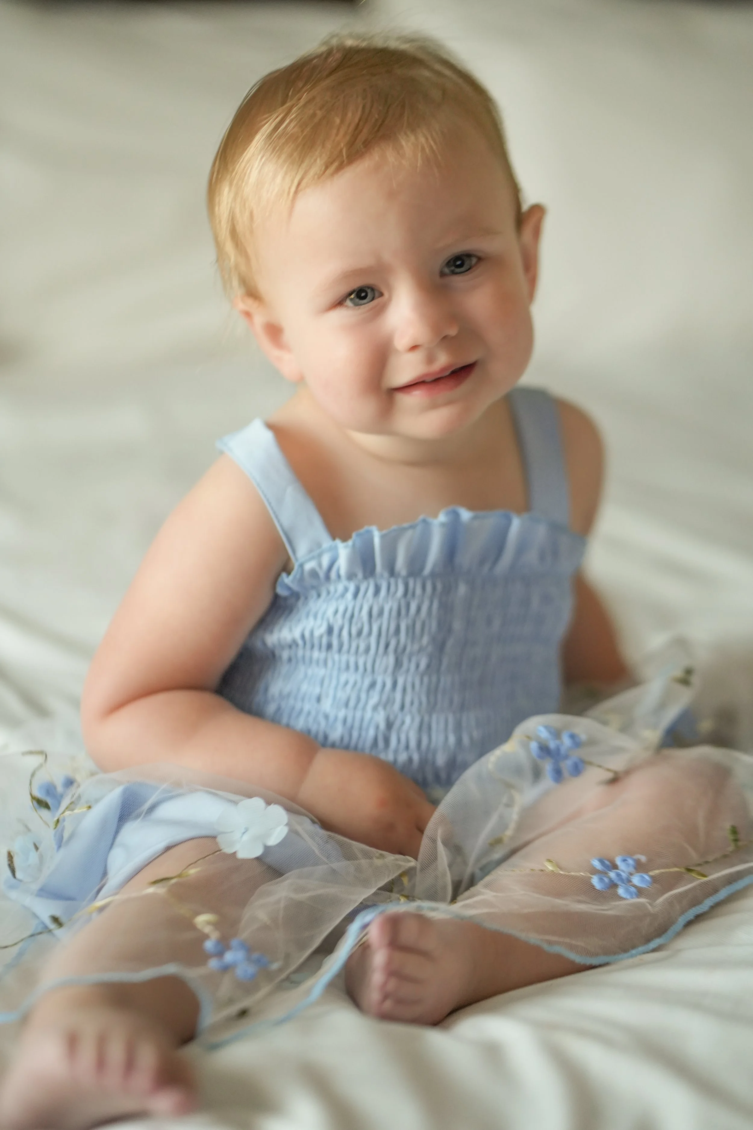 A young child with red hair and blue eyes sitting on a bed, wearing a light blue dress with ruffled straps, surrounded by sheer floral fabric with small blue flowers, smiling and looking at the camera.