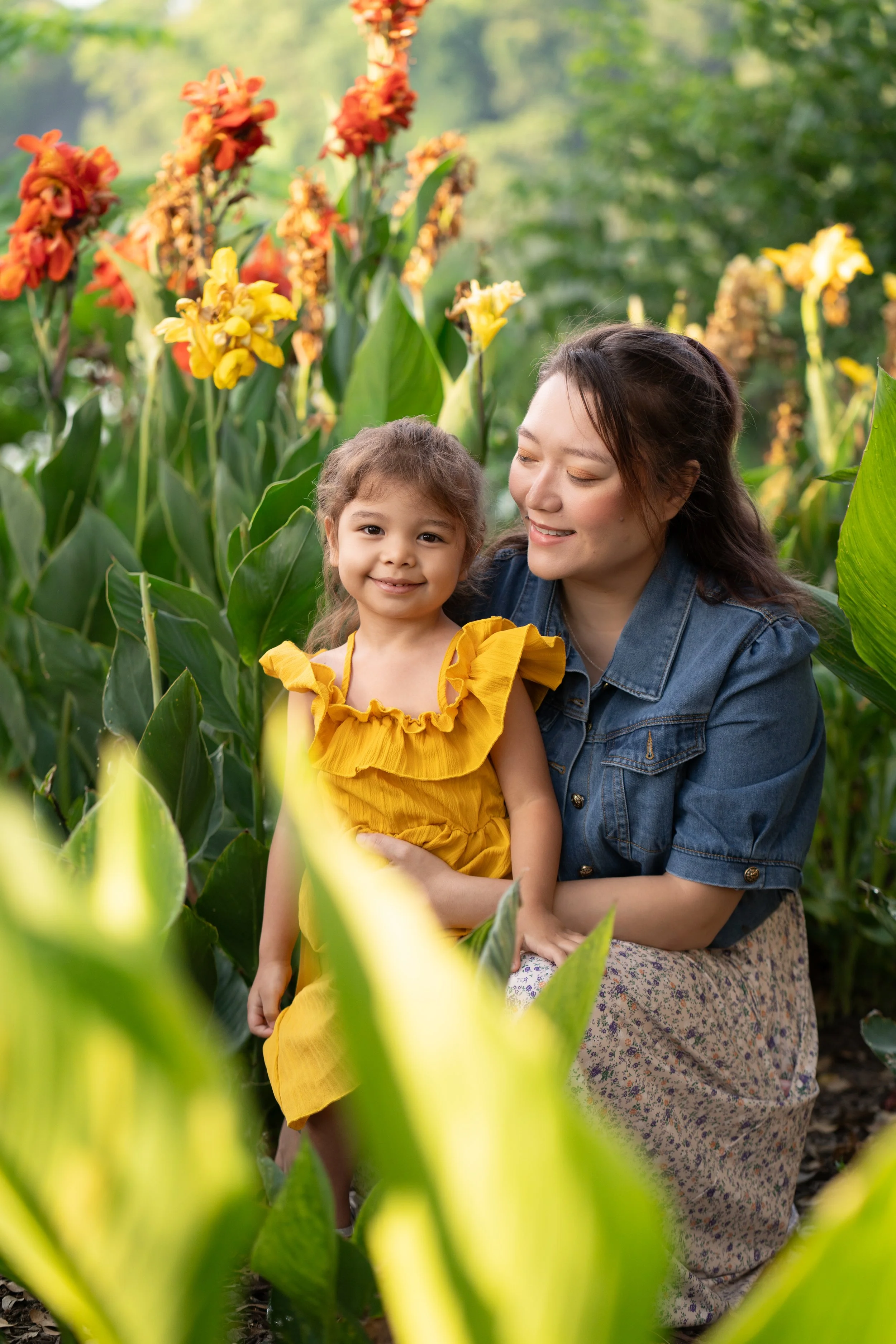 A young girl in a yellow dress and a woman, possibly her mother, are smiling and embracing each other among tall green plants with yellow and orange flowers, in a garden or park setting.