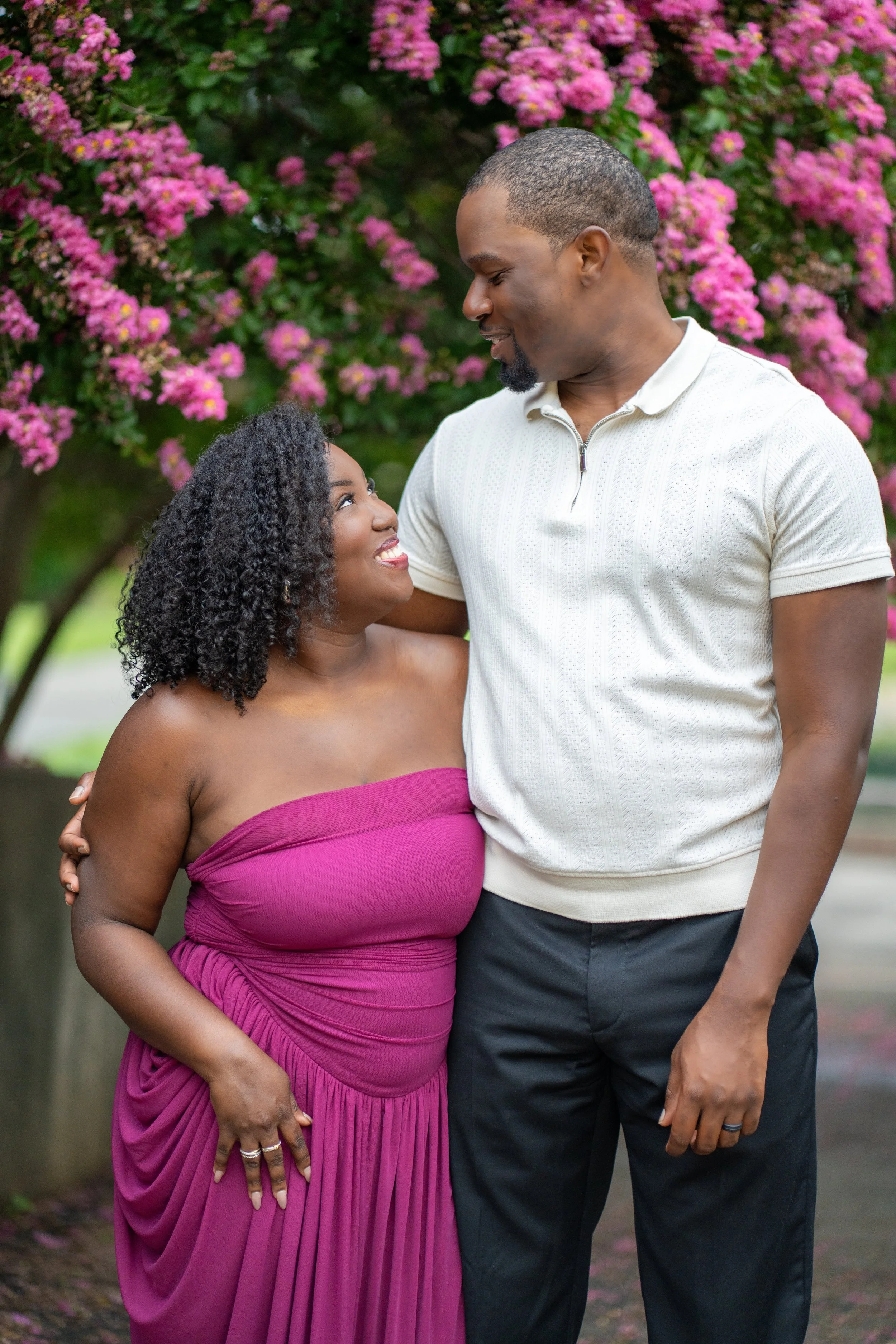 An African American woman in a strapless magenta dress, standing close and smiling up at an African American man in a white zip-up shirt, outdoors with pink flowers and greenery in the background.