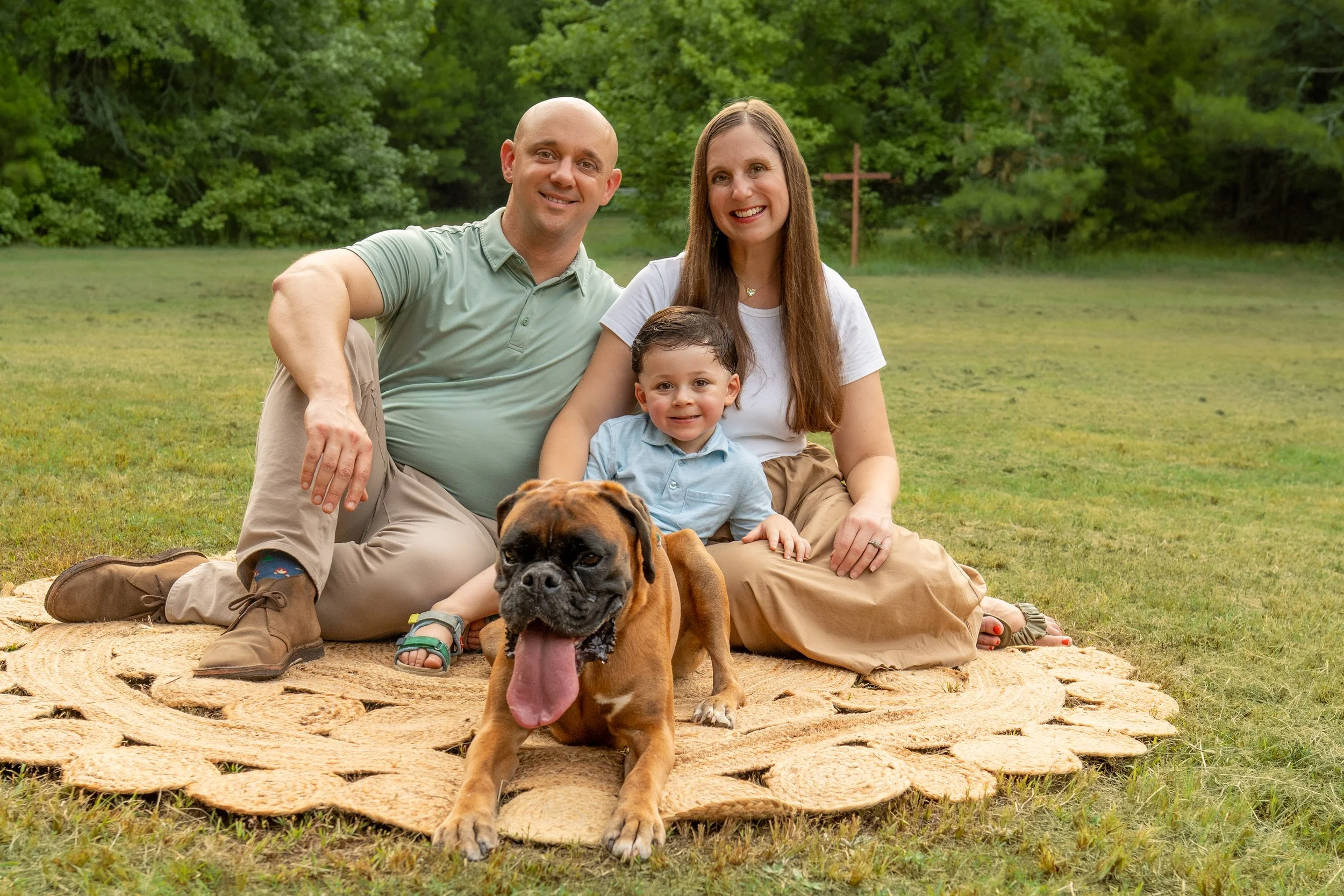 A happy family of three (a man, a woman, and a young boy) sitting on a blanket outdoors with their dog in a grassy, wooded area during daytime.
