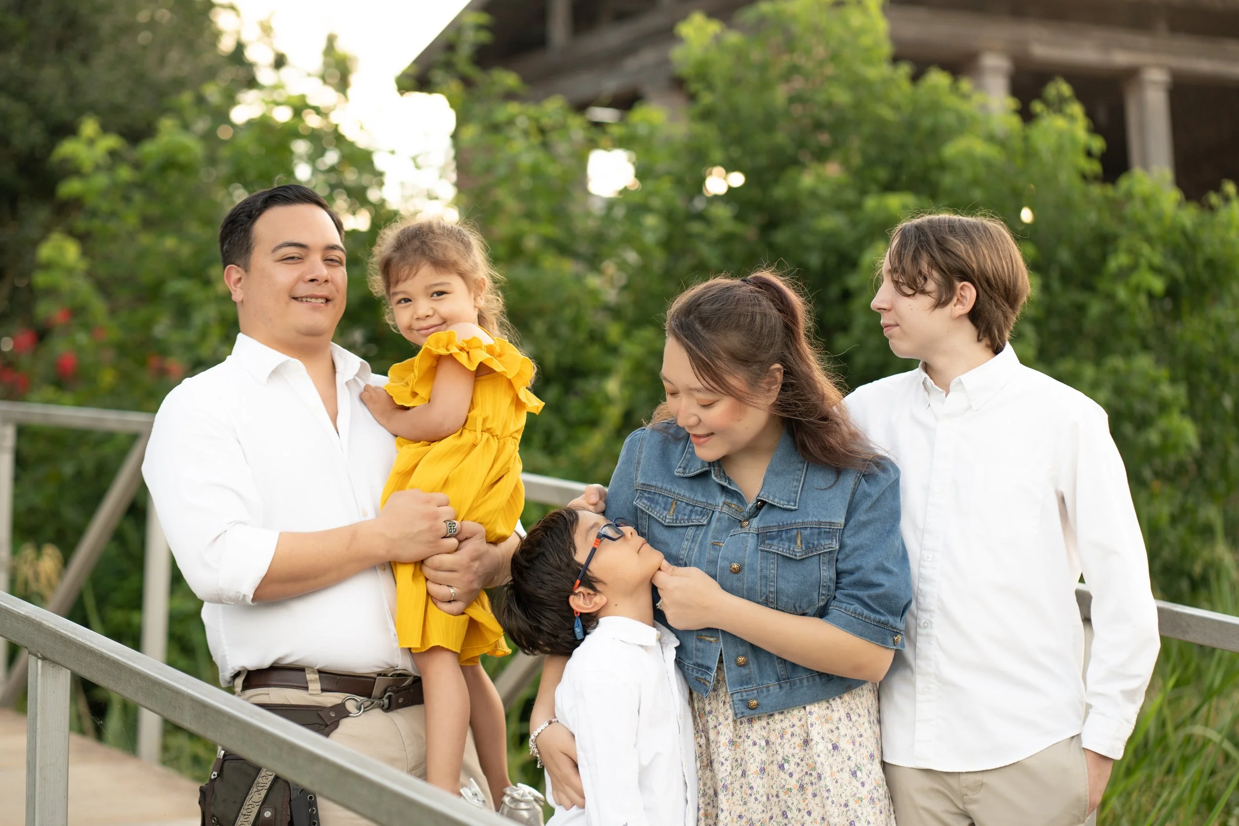 A group of five people, including a child being held by a smiling man, with greenery and a wooden structure in the background.