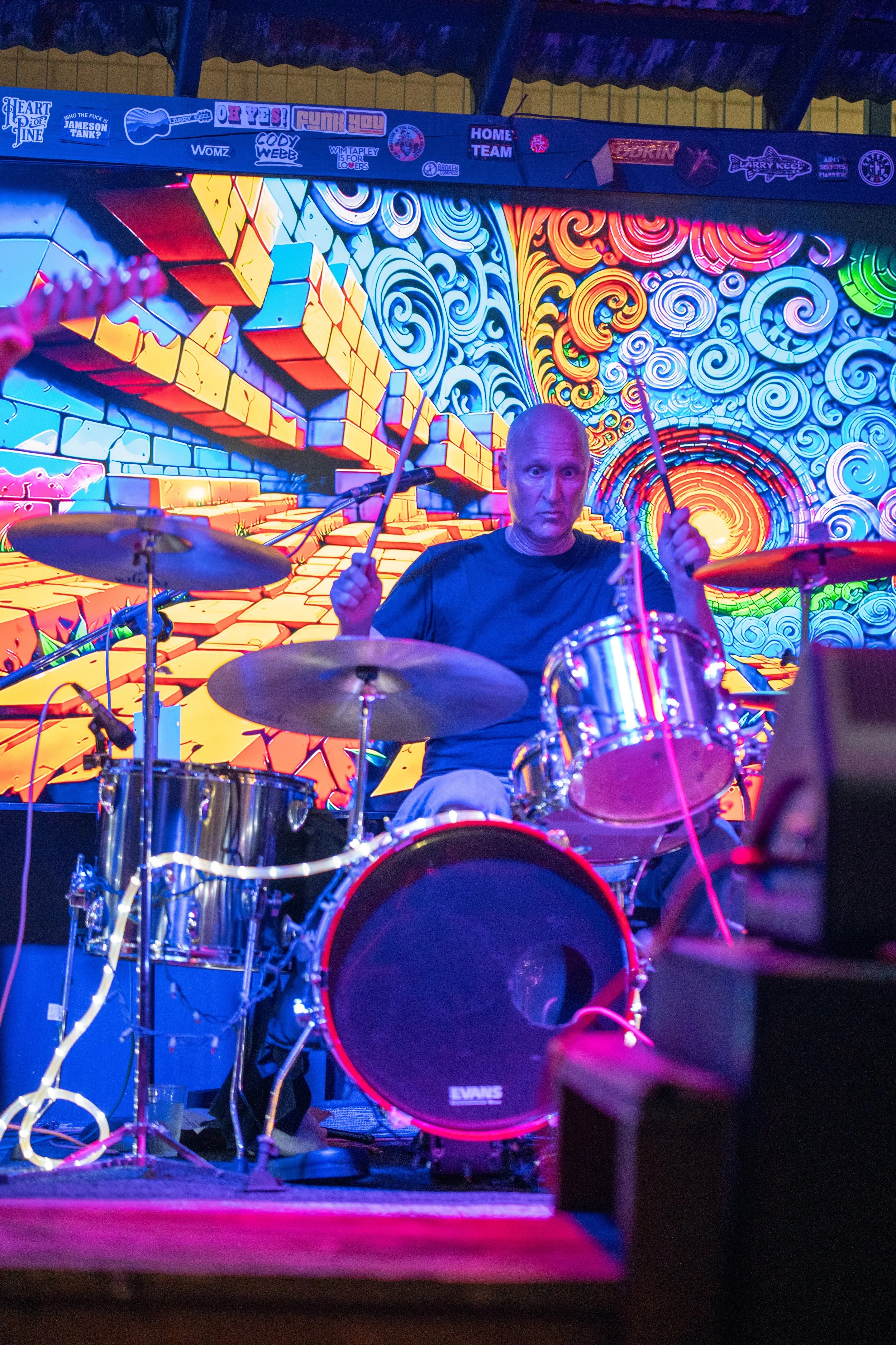 A drummer playing a drum set on stage with a vibrant, swirl-patterned background. The stage is decorated with colorful digital art and stickers on the wall.