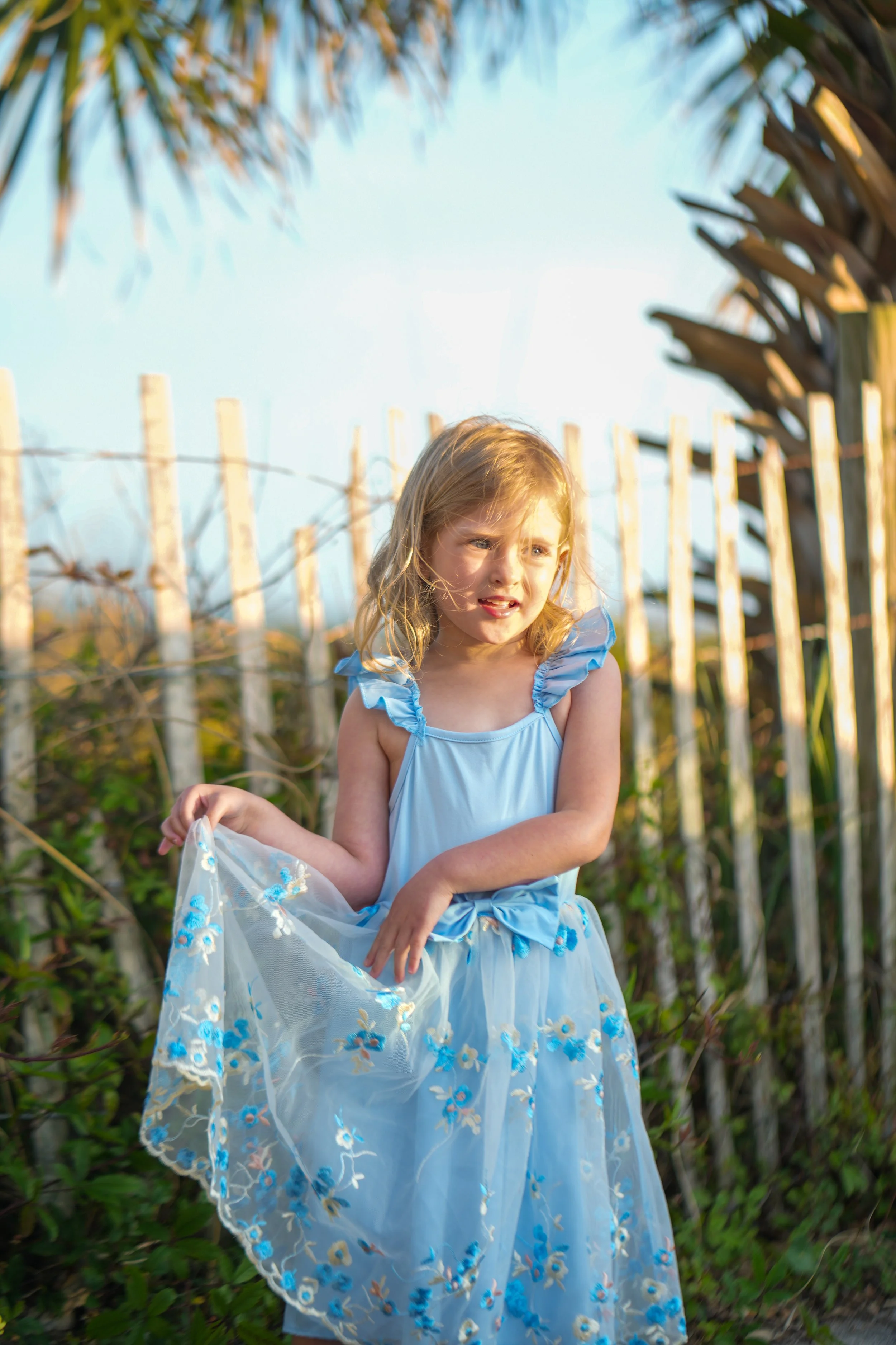 A young girl in a light blue dress with ruffled sleeves and a flowing skirt, holding a sheer fabric with embroidered blue flowers. She has blonde hair and a slightly distressed or focused expression. The background consists of a palm tree canopy and a wooden fence.
