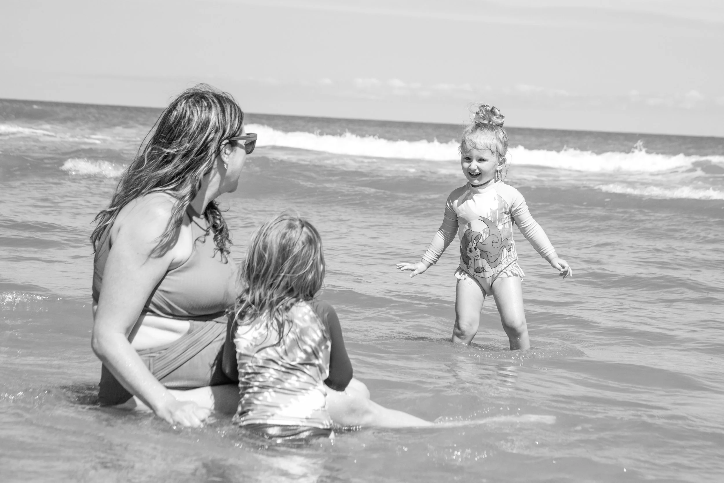 A woman, two children, and a girl playing in the ocean at the beach, with the girl jumping in the water.