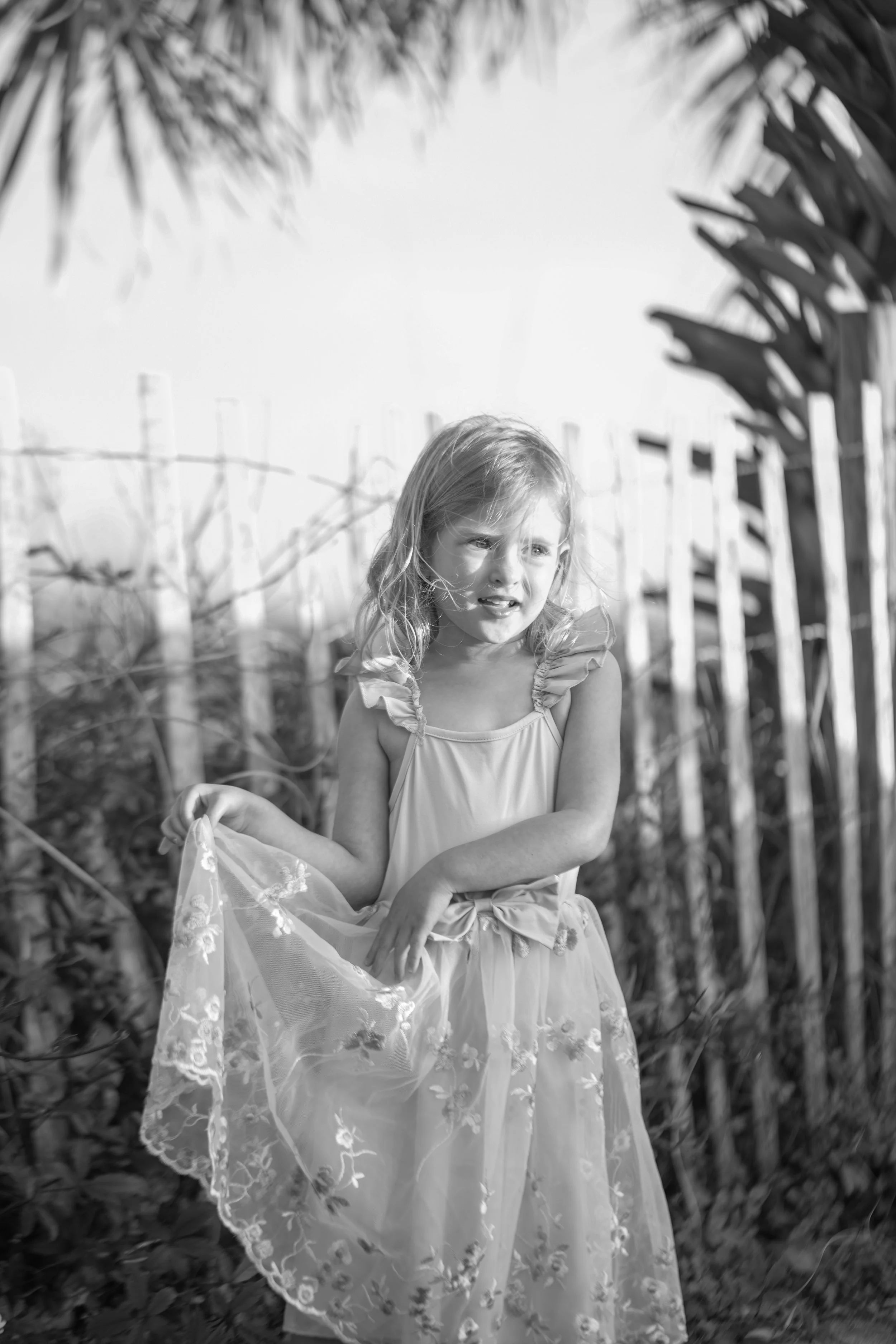 A young girl in a light dress with floral embroidery, sitting outdoors among tall plants and a wooden fence, with a distressed expression.