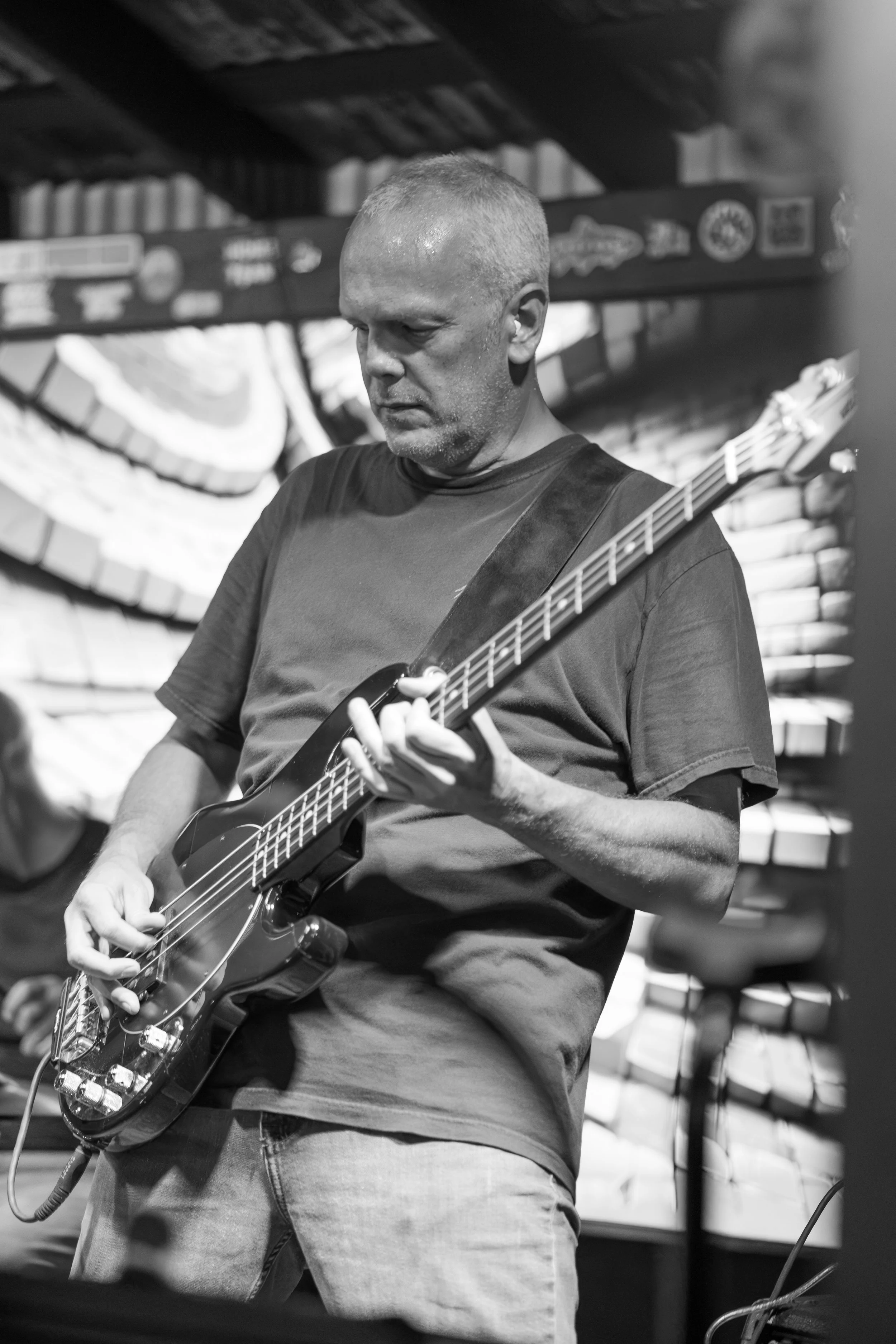A man with short hair and a black t-shirt playing an electric guitar in a room with a curved wooden ceiling and various posters or signs on the wall.