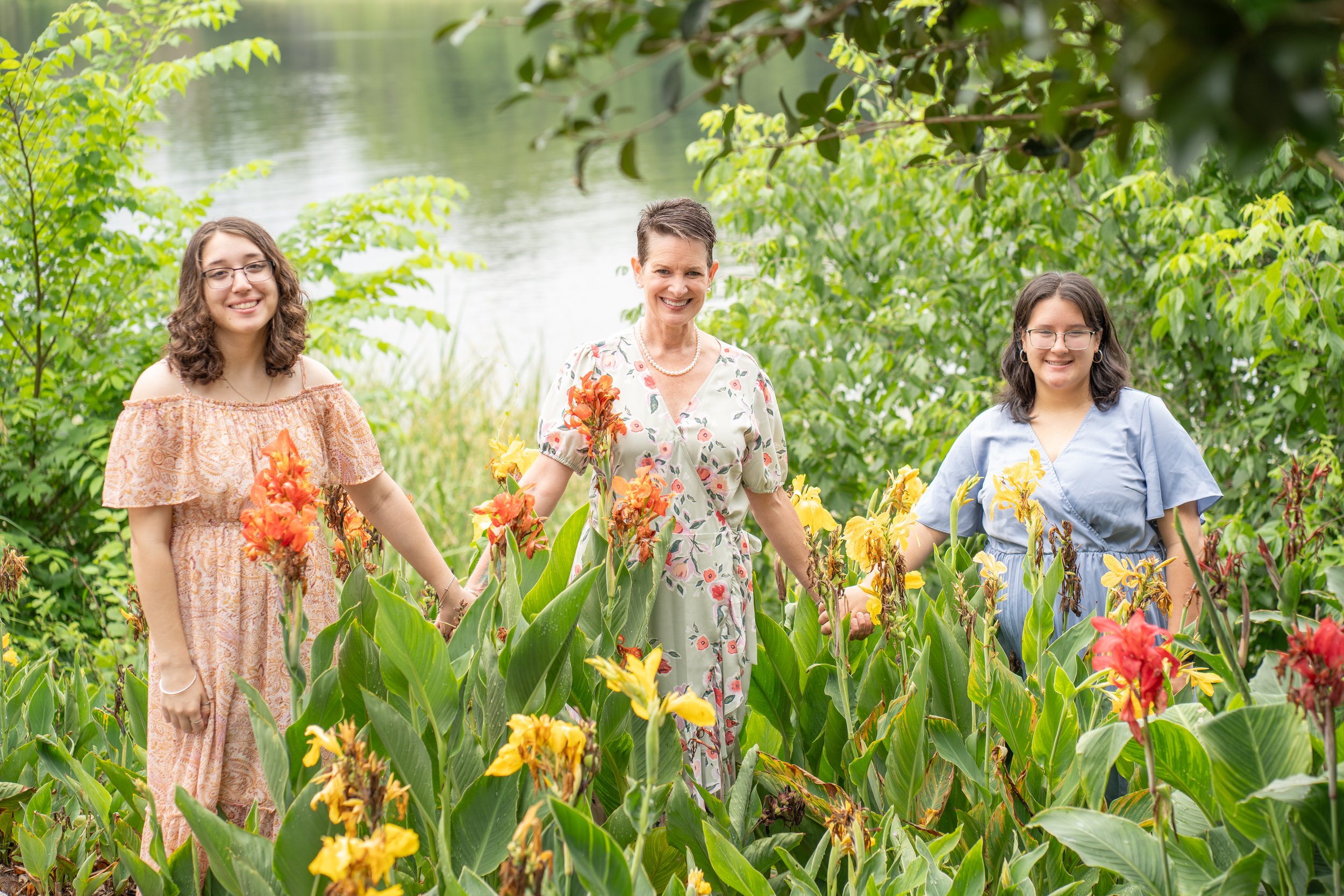 Three women, likely a grandmother and two granddaughters, holding hands and standing in a lush garden with blooming flowers near a body of water, on a cloudy day.
