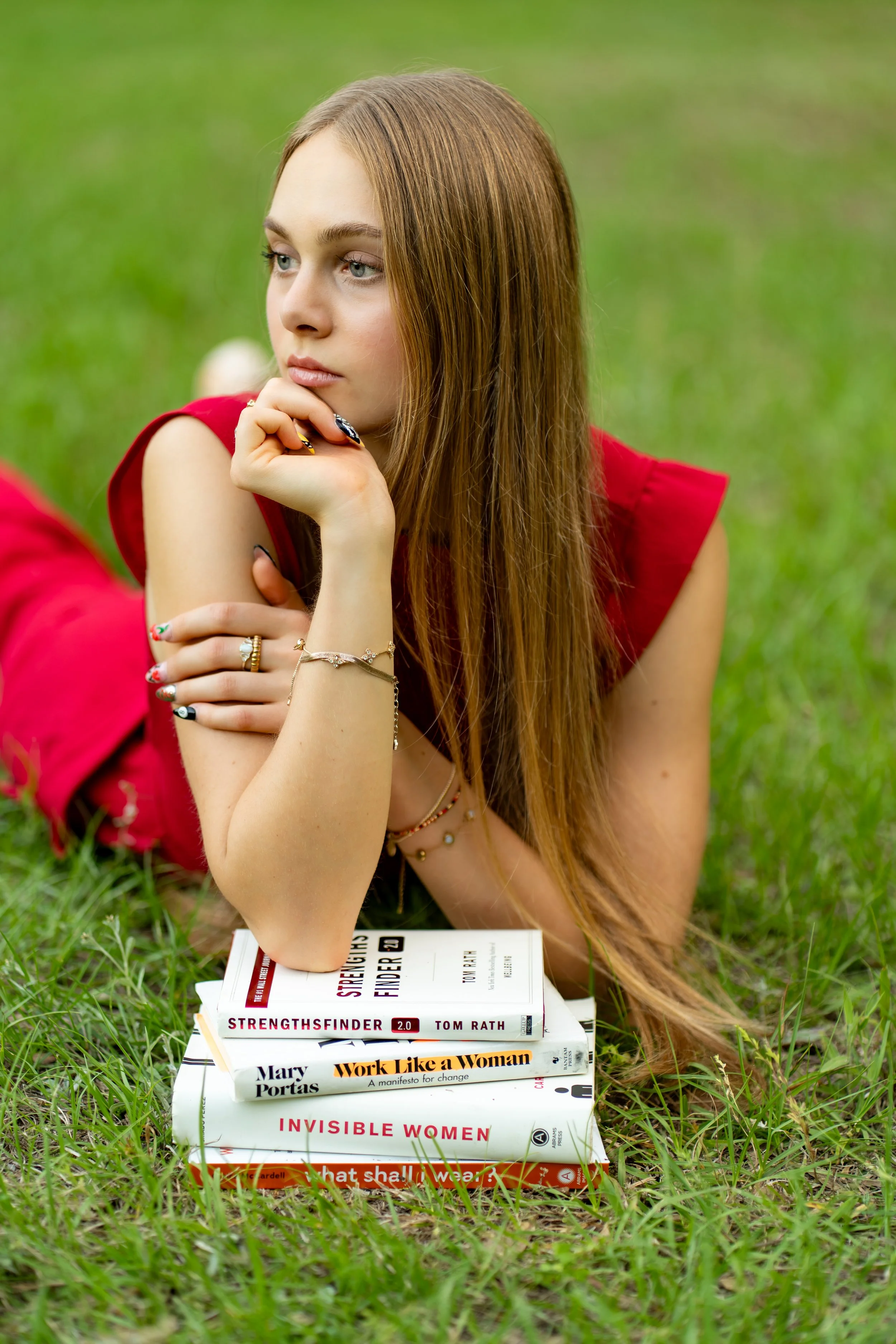 A young woman with long reddish-brown hair is lying on the grass outdoors, resting her chin on her hand. She is wearing a red sleeveless top and multiple rings and bracelets. In front of her, there are several books stacked on the grass, including titles like "StrengthsFinder," "Work Like a Woman," "Invisible Women," and others.