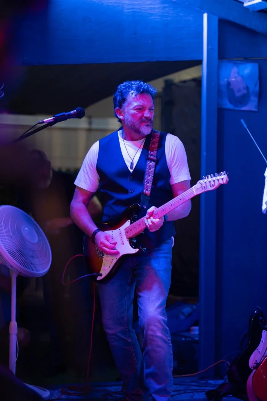A man with a beard and short curly hair playing an electric guitar in a dimly lit venue with purple and blue lighting, wearing a white t-shirt, a dark vest, and jeans.