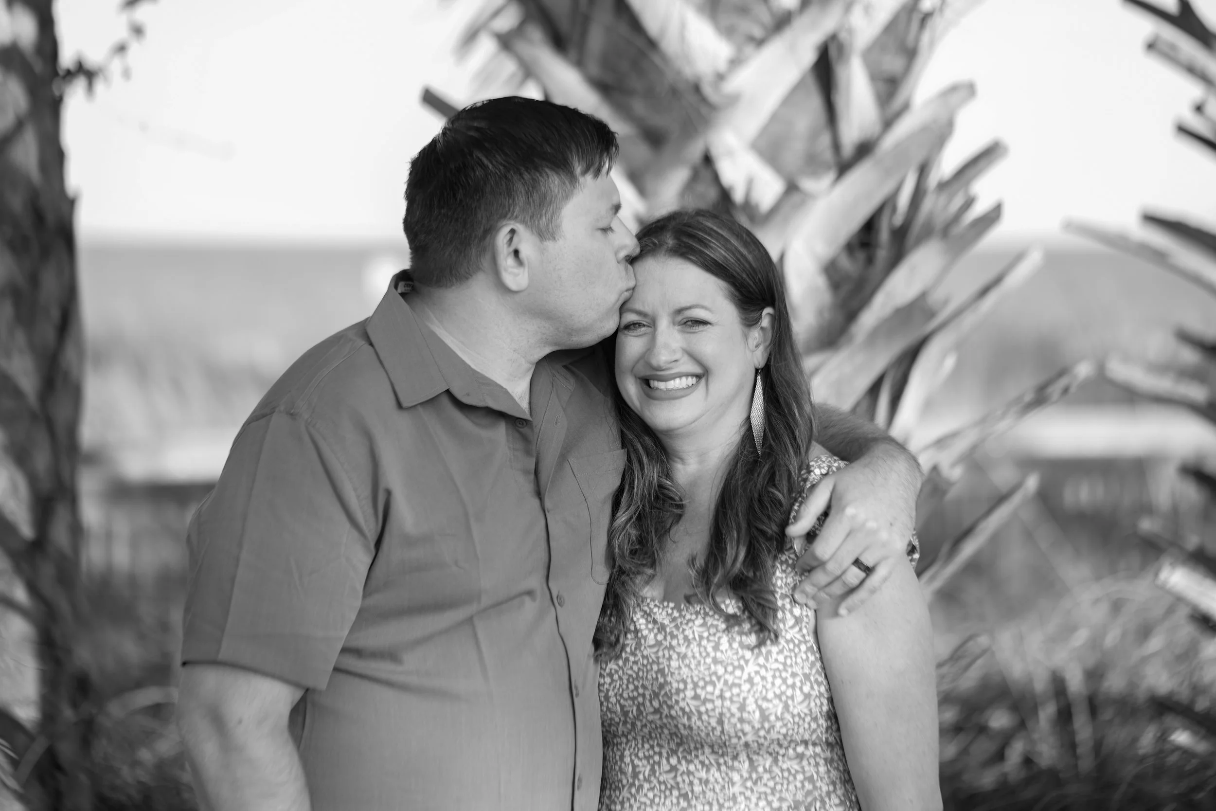A black and white photo of a man kissing a woman on the forehead, both smiling and embracing each other in front of a large plant with broad leaves.