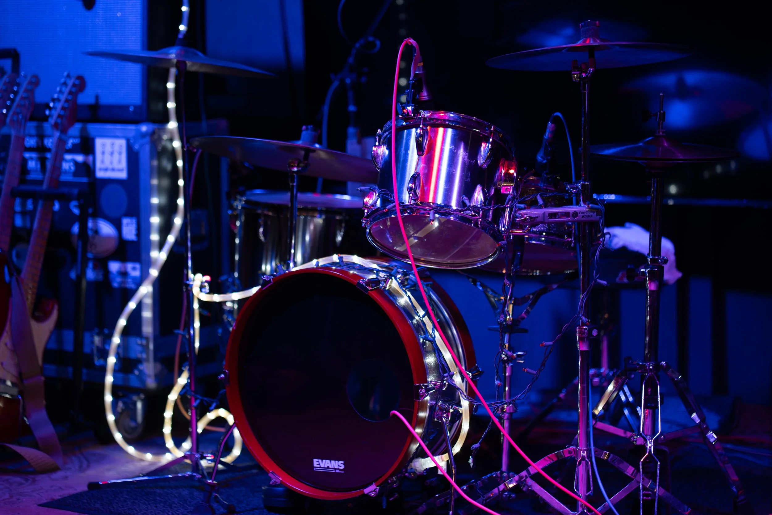 A drum set with a shiny metallic finish, illuminated by purple and blue stage lights, with guitars and music equipment in the background.