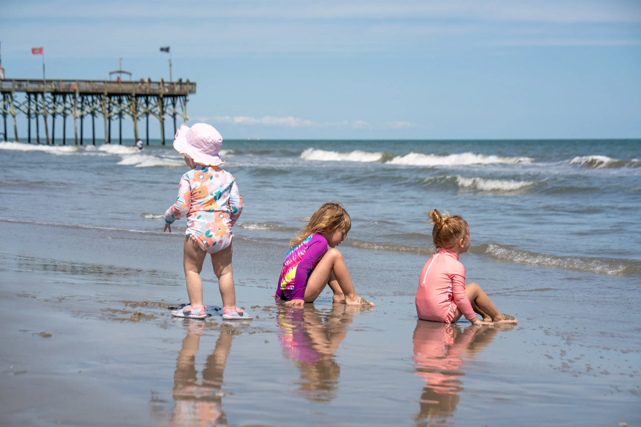 Four young children, three girls and one boy, playing at the beach near the shoreline, with a pier in the background and waves crashing on the shore.