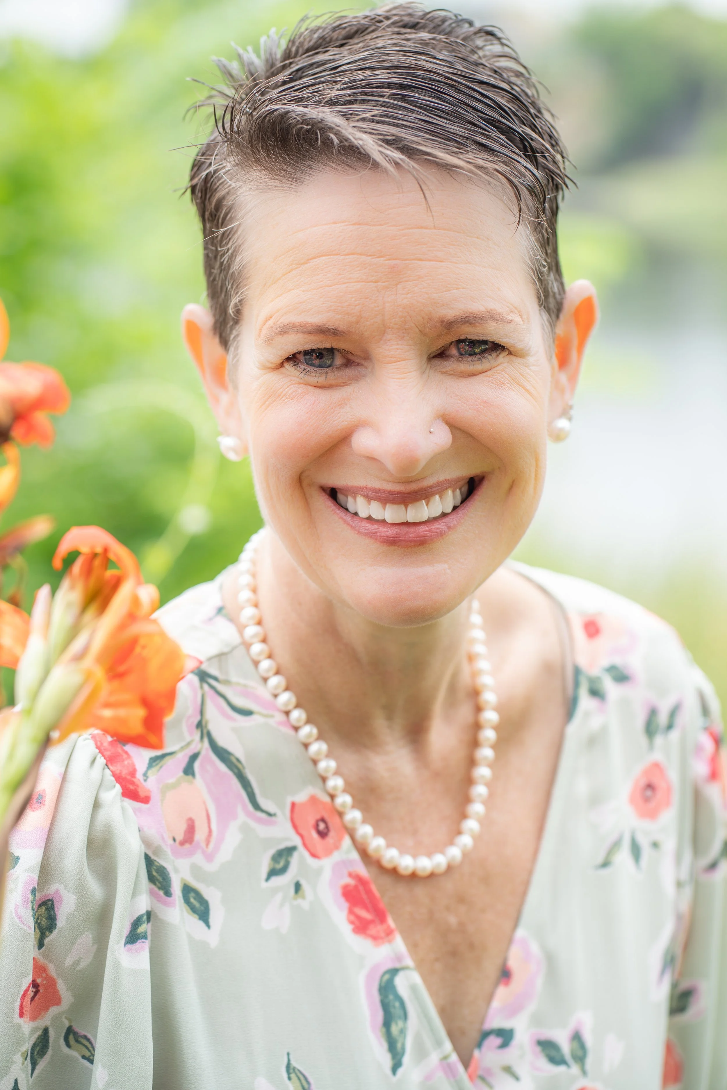 Close-up of a smiling woman with short, dark, wet hair wearing pearl earrings and a floral dress, outdoors with green blurred background.