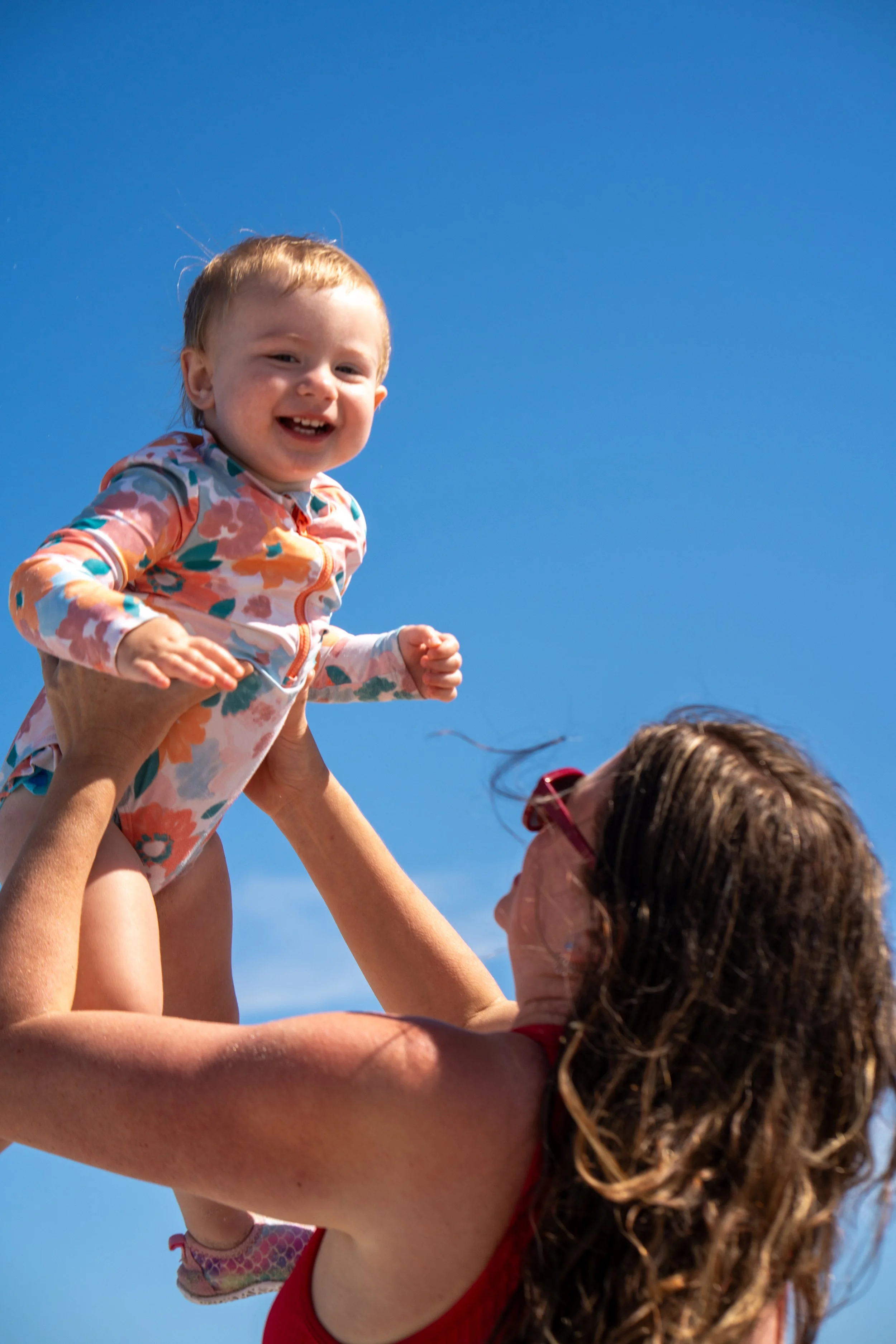 A smiling young girl with light brown hair being raised into the air by an adult, against a clear blue sky.