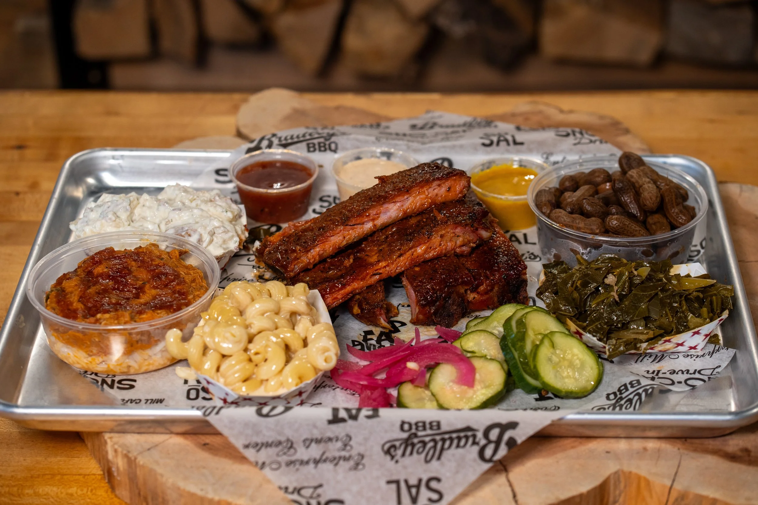 A metal tray with barbecue food including sliced smoked ribs, potato salad, baked beans, wilted greens, pickles, chopped onions, and assorted sauces in small cups, arranged on decorative paper packaging.