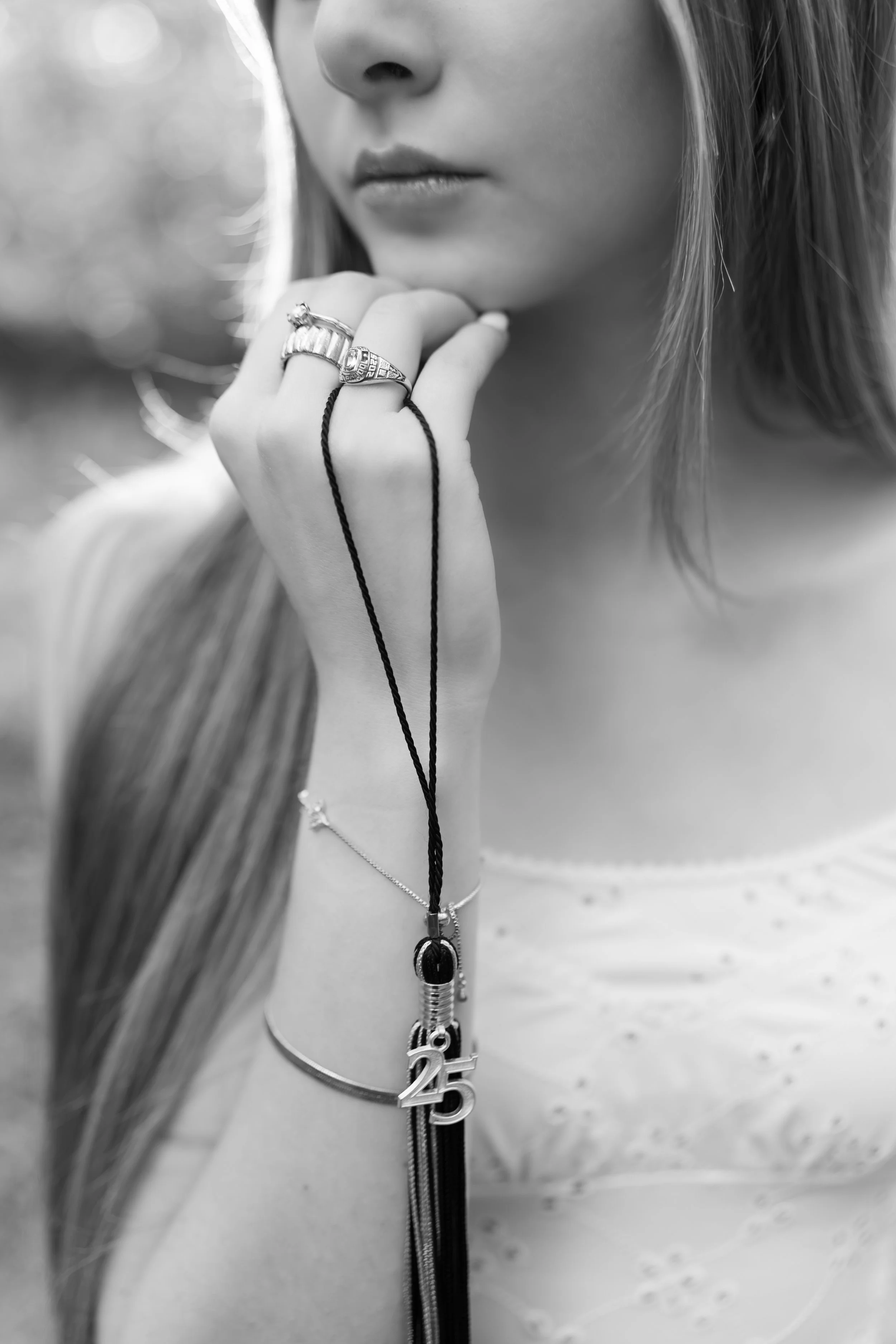 A close-up of a woman's face and hand, with necklaces and rings, holding a black necklace with a pendant, wearing a zippered top, in black and white.